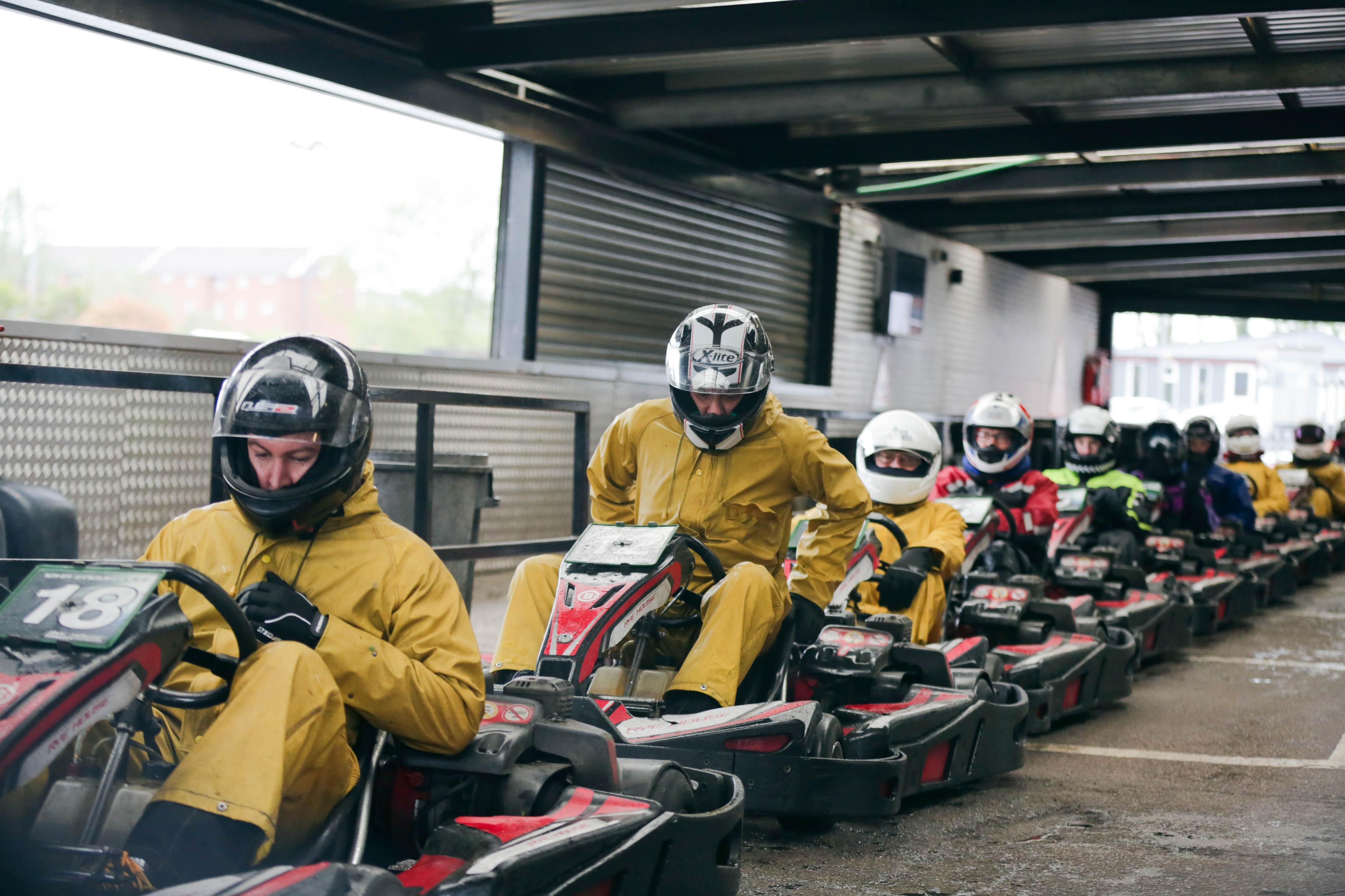 Line of go-kart drivers in yellow suits and helmets preparing to race indoors, with garage windows and metal barriers visible.