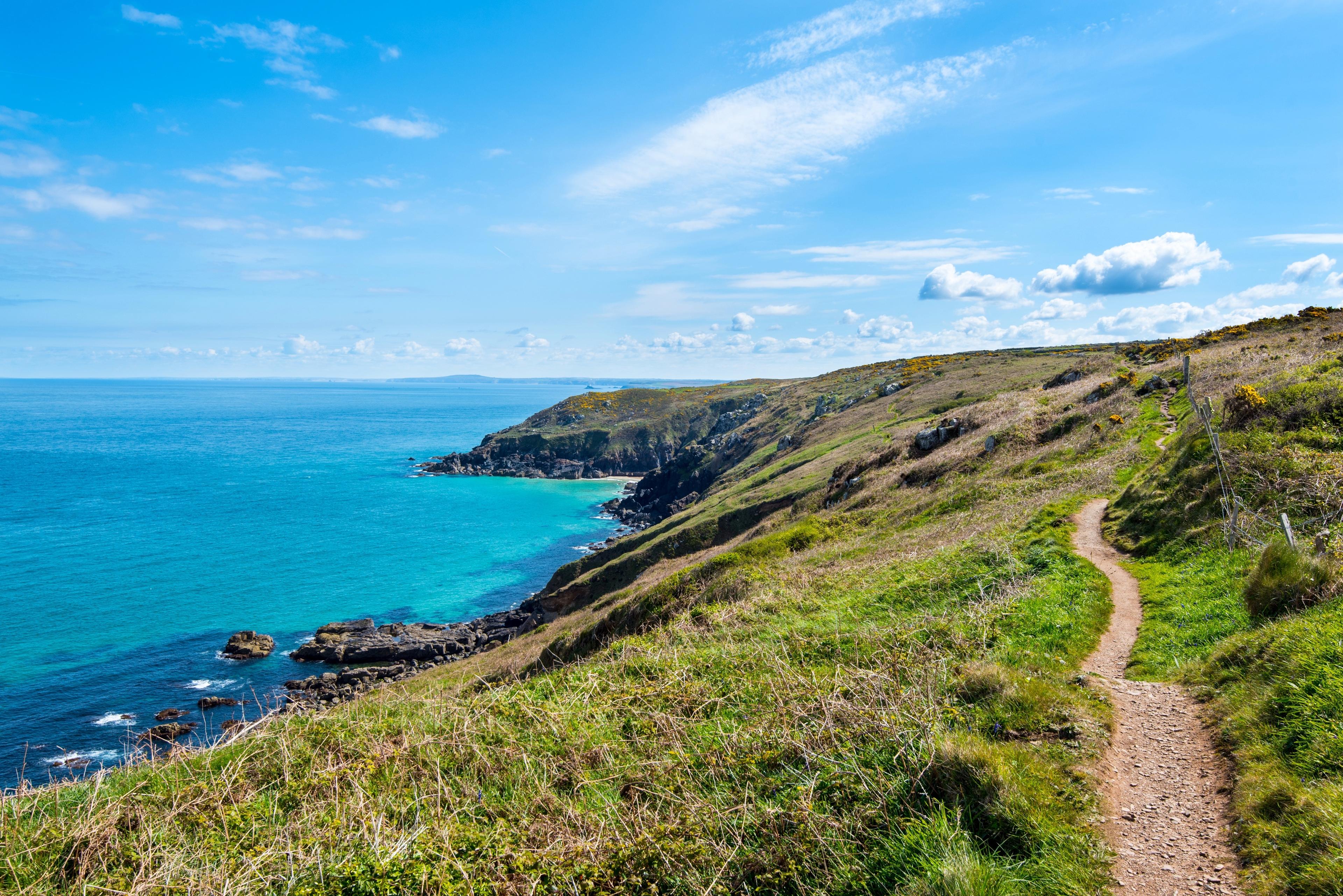 Coastal landscape with a dirt path along grassy cliffs overlooking clear blue sea under a bright sky with scattered clouds.