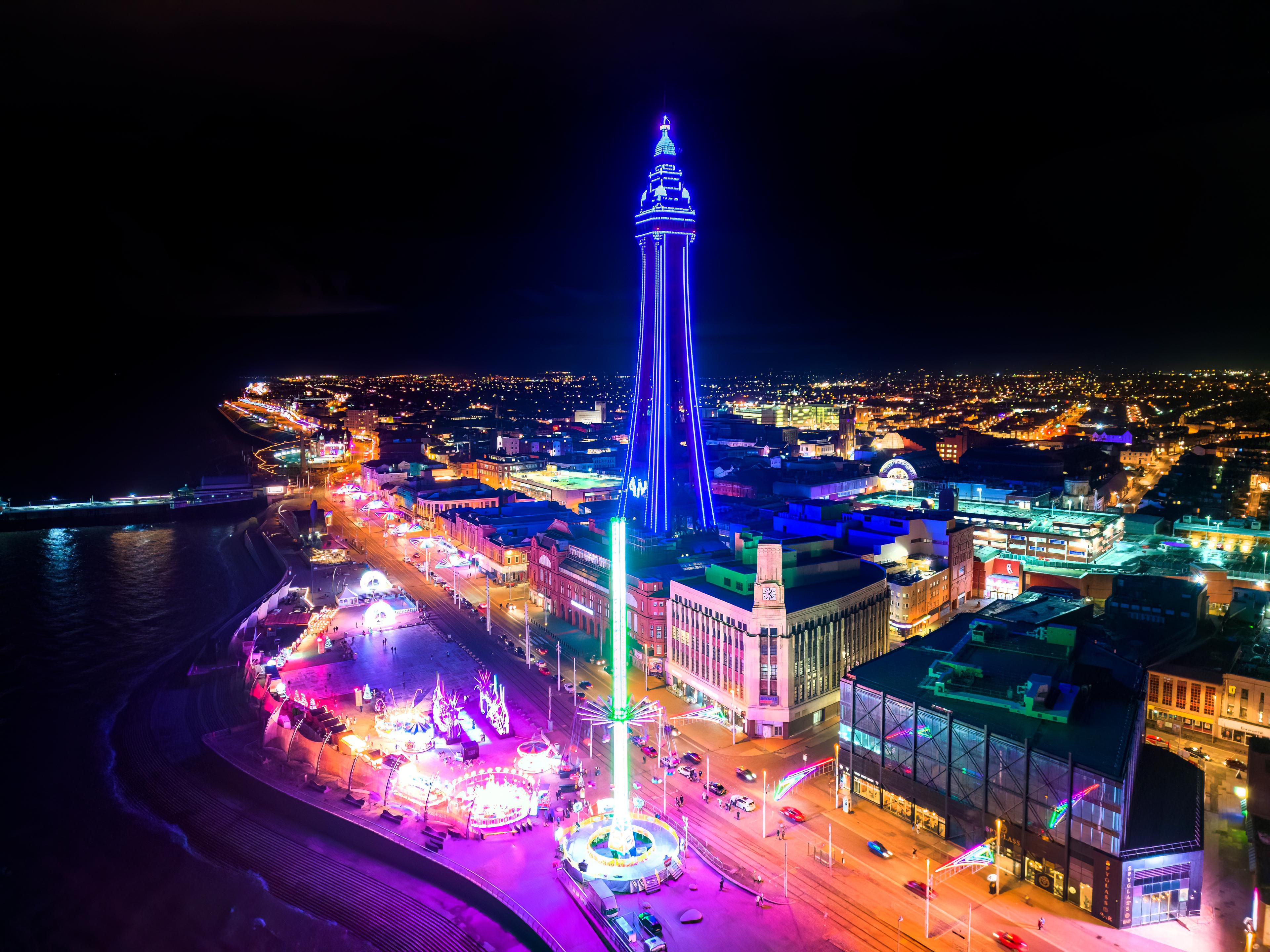 A vibrant nighttime aerial view of a cityscape with a brightly lit tower and colorful lights along the streets and beachfront.