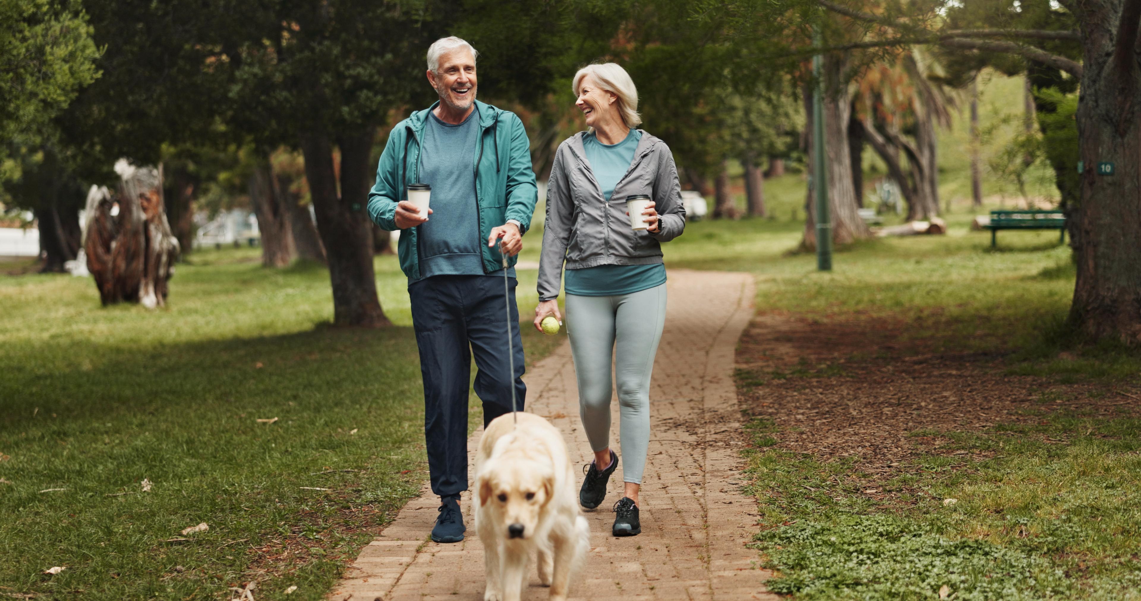 Older couple walking their dog in a park, holding coffee cups, and smiling at each other amidst lush greenery.