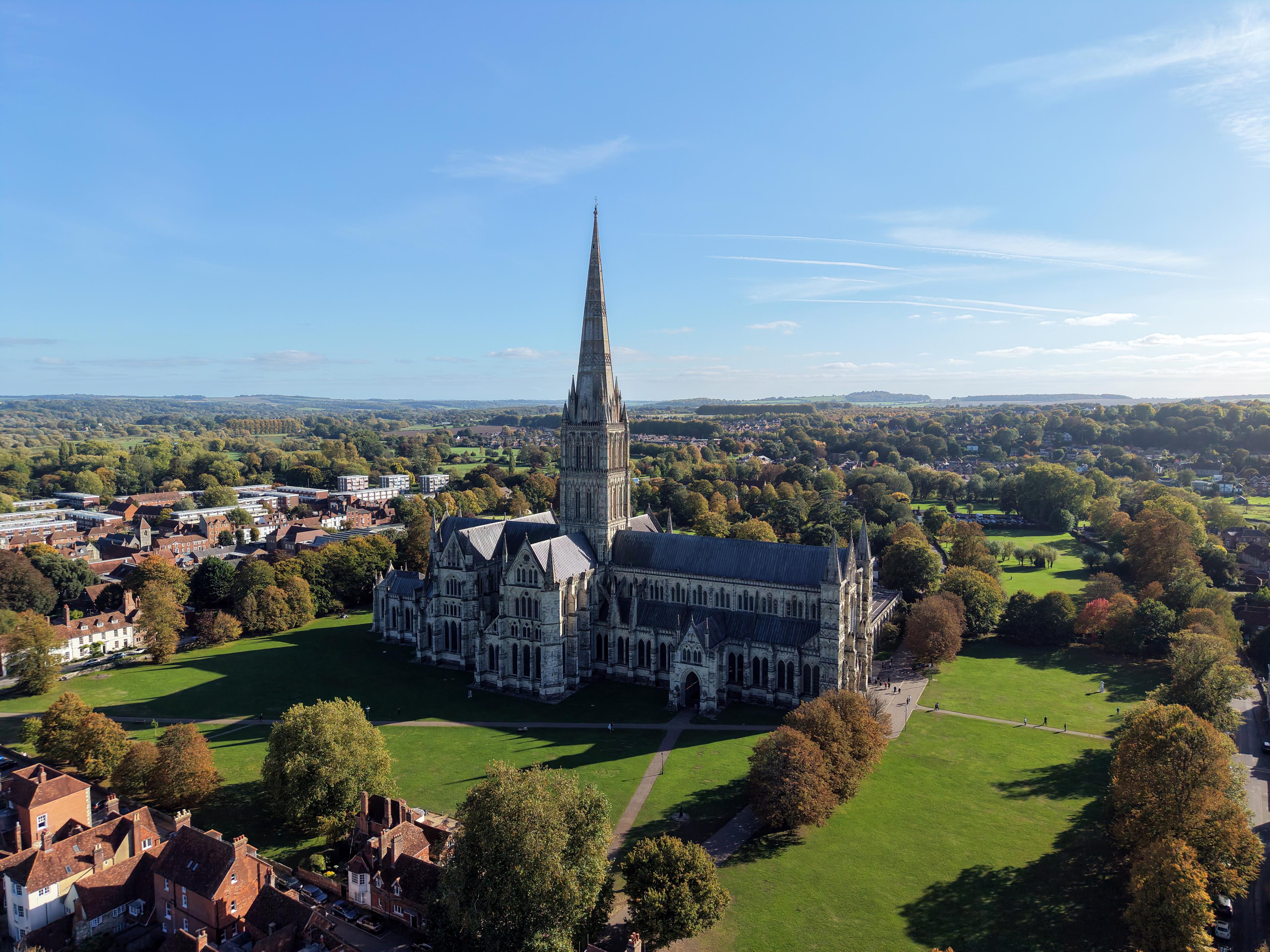 Aerial view of a Gothic cathedral with a tall spire surrounded by greenery and city buildings under a clear blue sky.