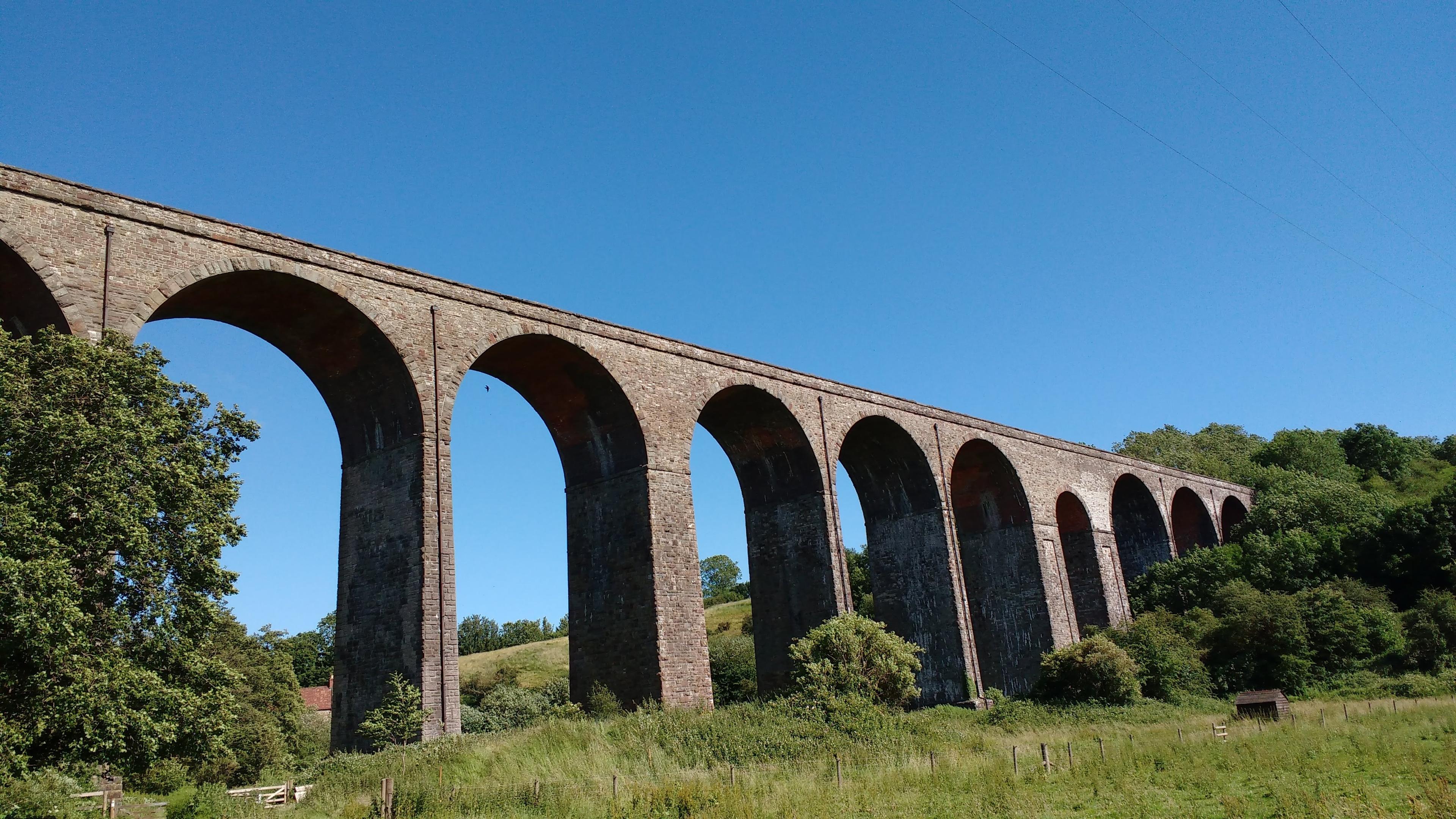 Stone viaduct with multiple arches spanning over a lush, green landscape under a clear blue sky.