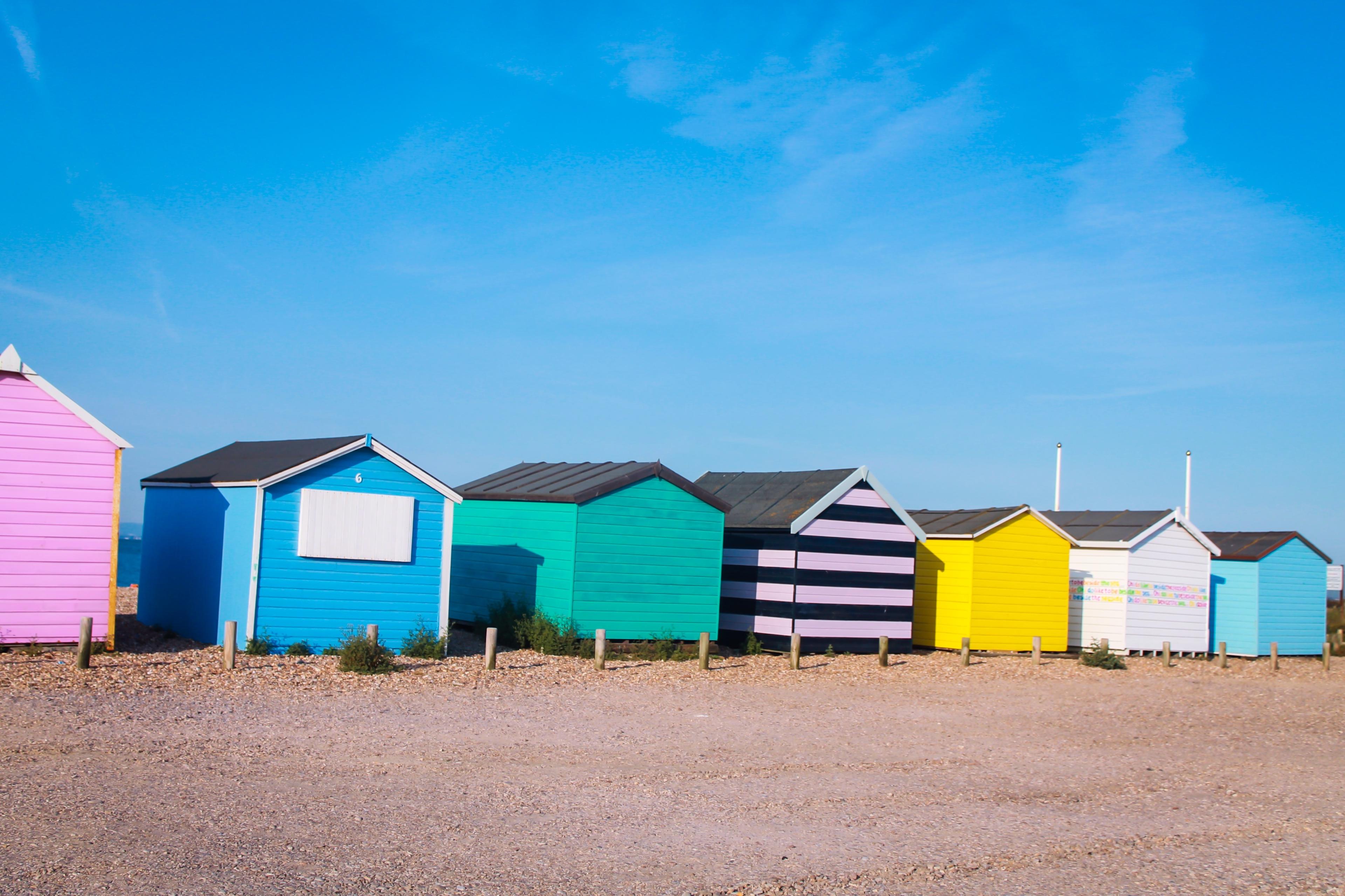 Colorful seaside beach huts in pink, blue, turquoise, striped purple, yellow, and white line a pebbly shore beneath a bright blue sky.