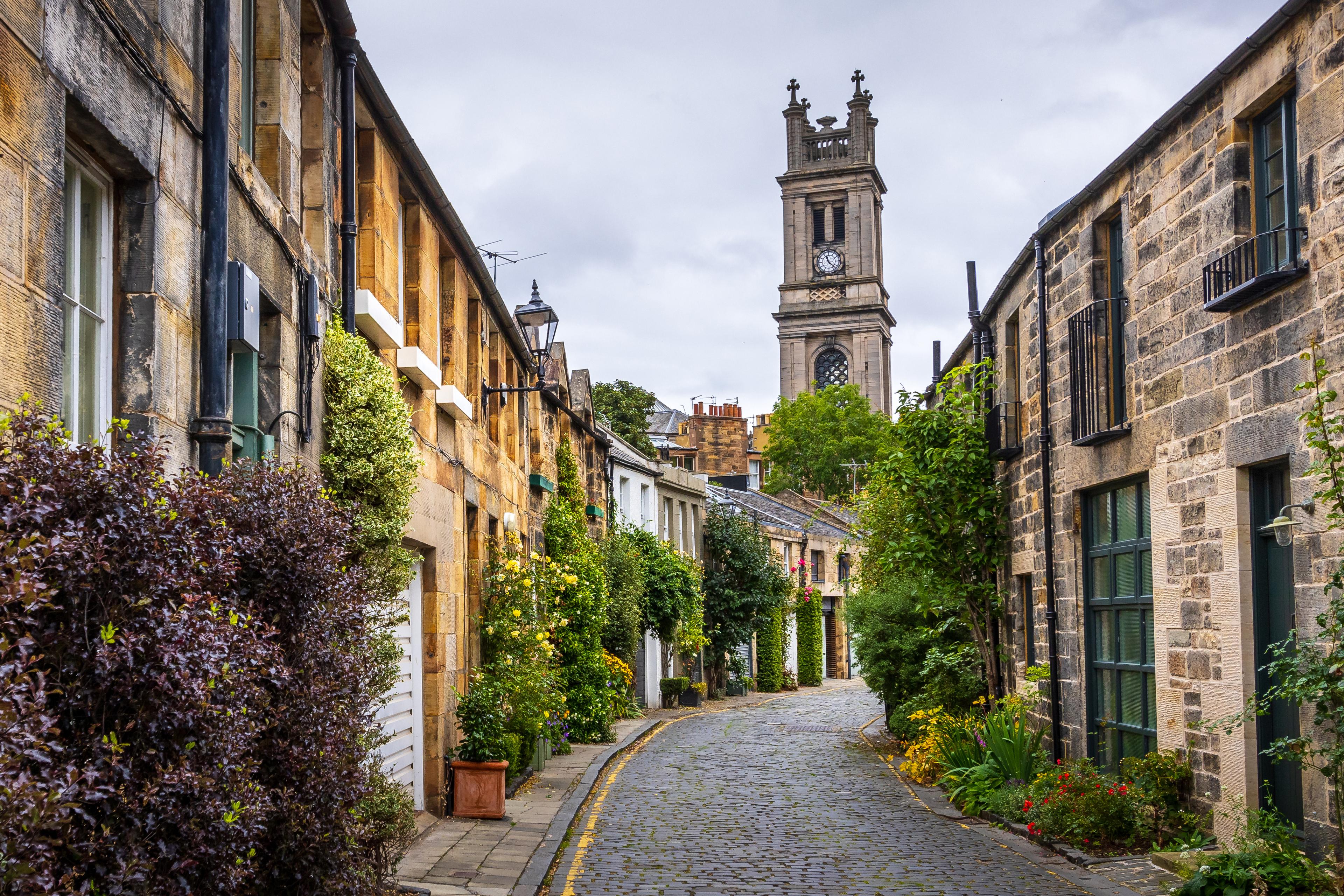 A narrow cobblestone street lined with charming stone buildings, lush greenery, and a clock tower in the background under a cloudy sky.