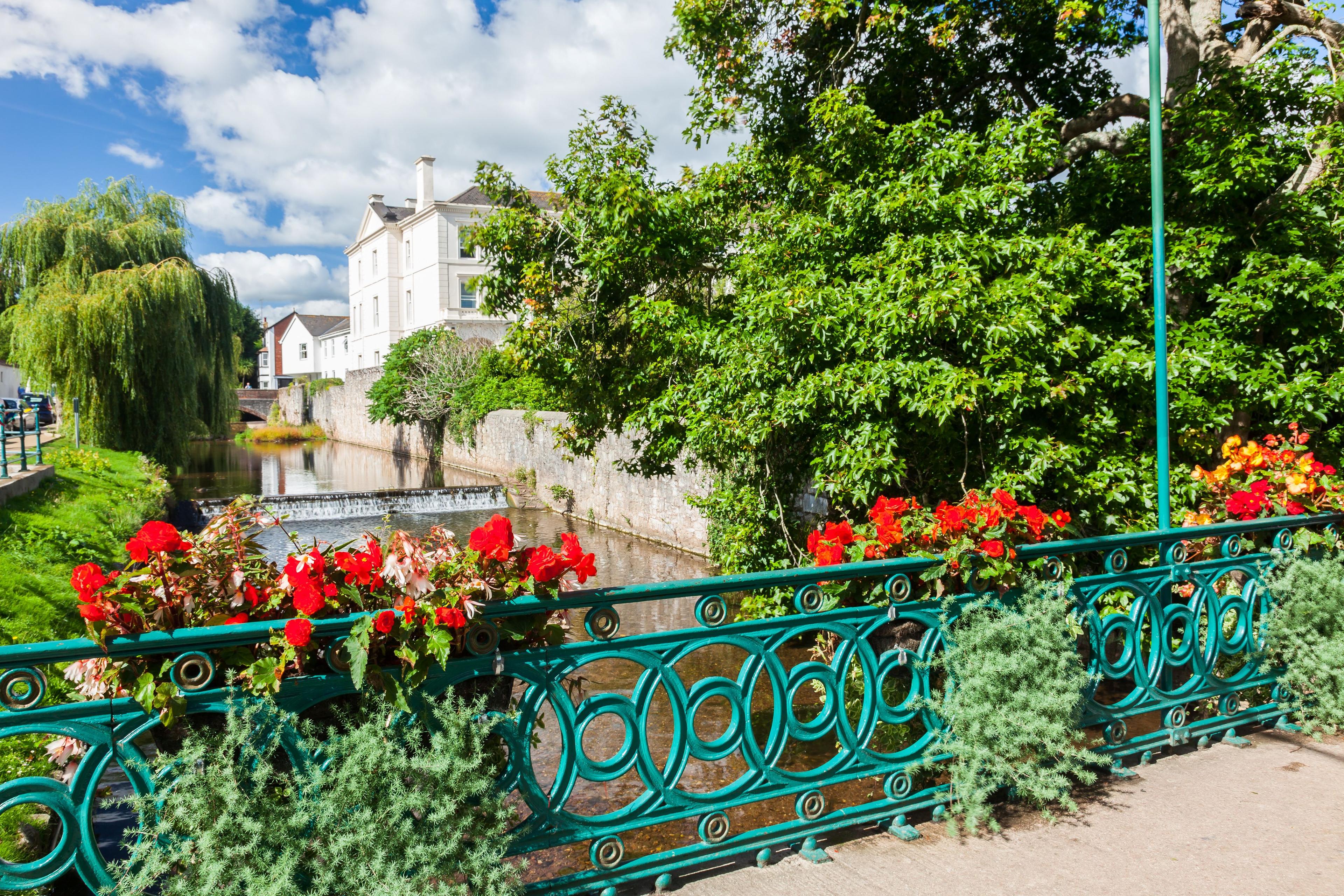 Bright riverside garden with red flowers, ornate teal bridge railing, leafy trees, willow, white houses, and blue sky; peaceful summer scene.