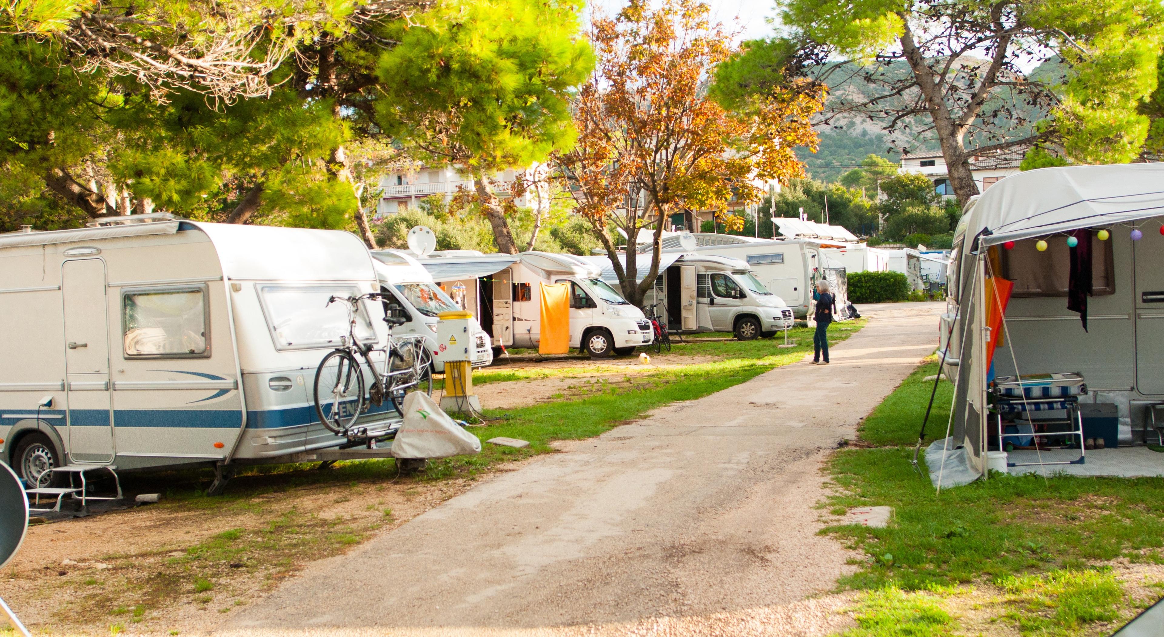 A campground with parked caravans and motorhomes on a sunny day, surrounded by trees and a narrow gravel path.
