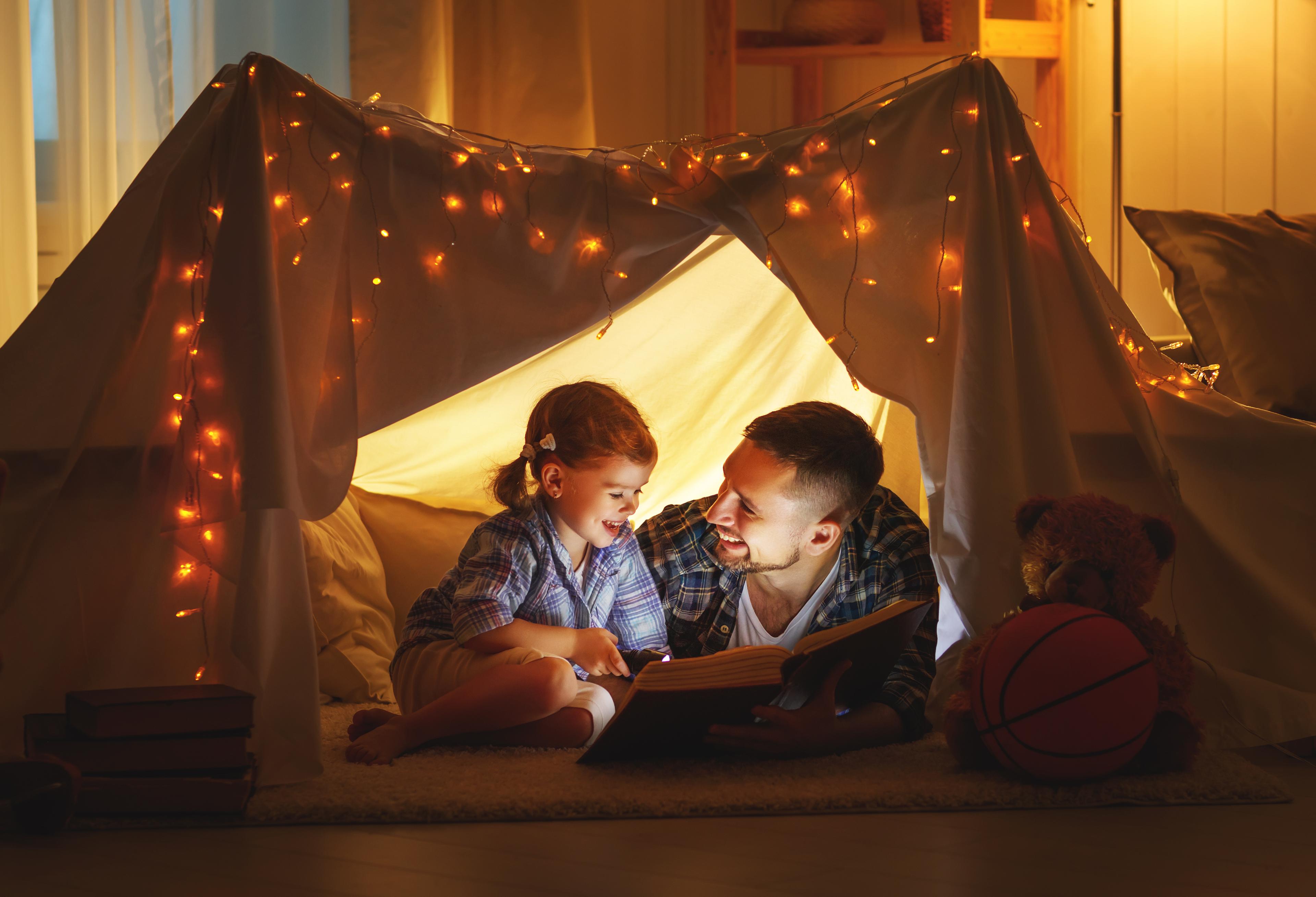A father and young daughter sit together inside a cozy blanket fort, reading a book by the warm glow of string lights. A teddy bear and basketball rest nearby.