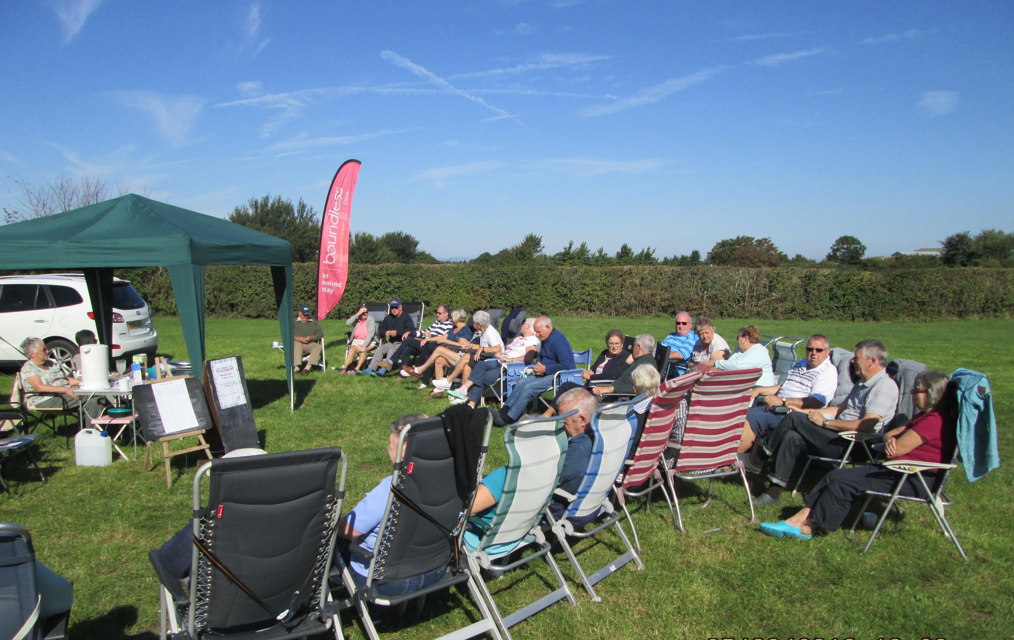 Group of people seated on lawn chairs in a sunny field near a green canopy