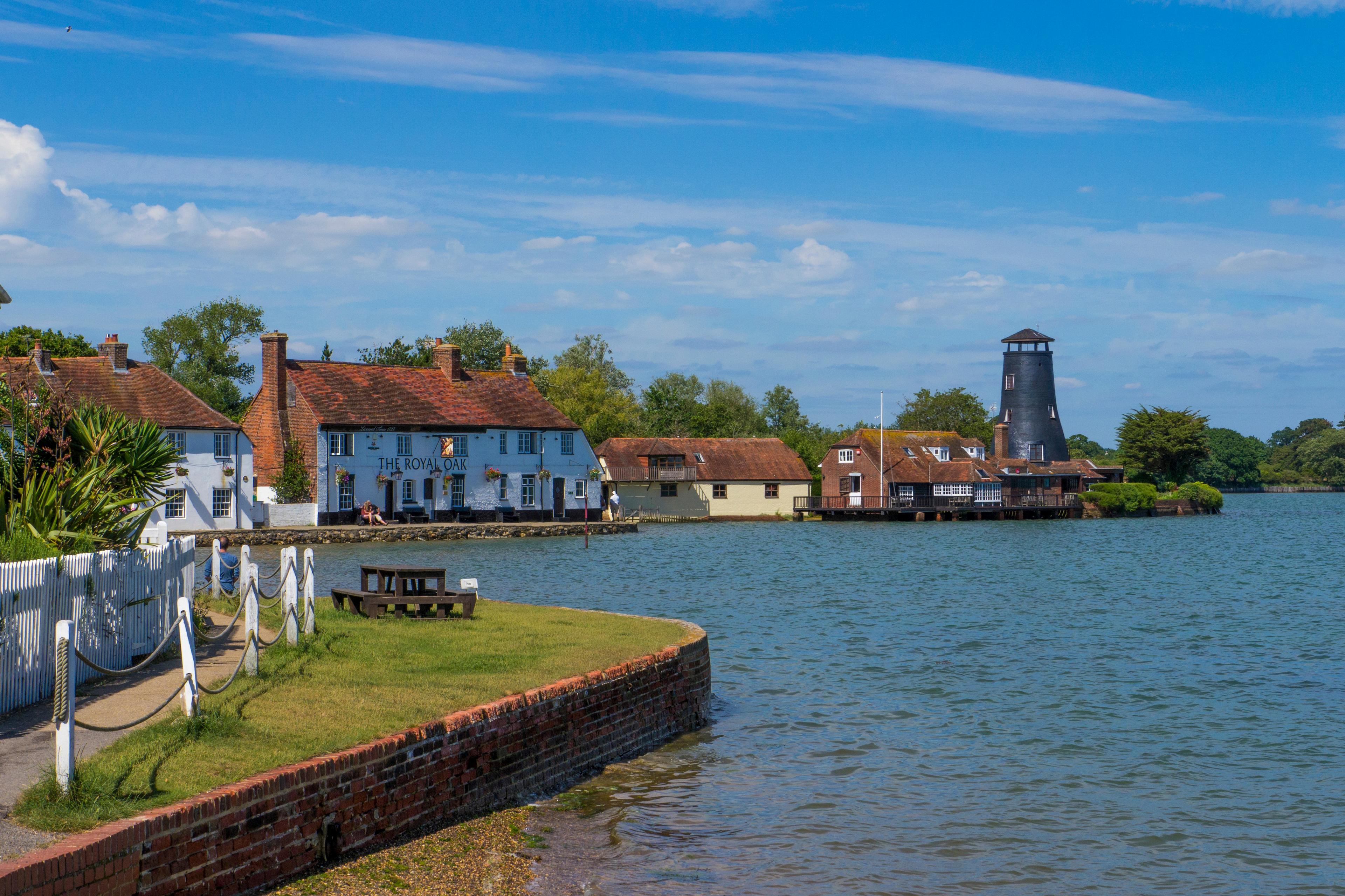 Scenic waterfront village with white cottages, red-tiled roofs, and a dark brick tower beside calm blue water under a bright, partly cloudy sky.