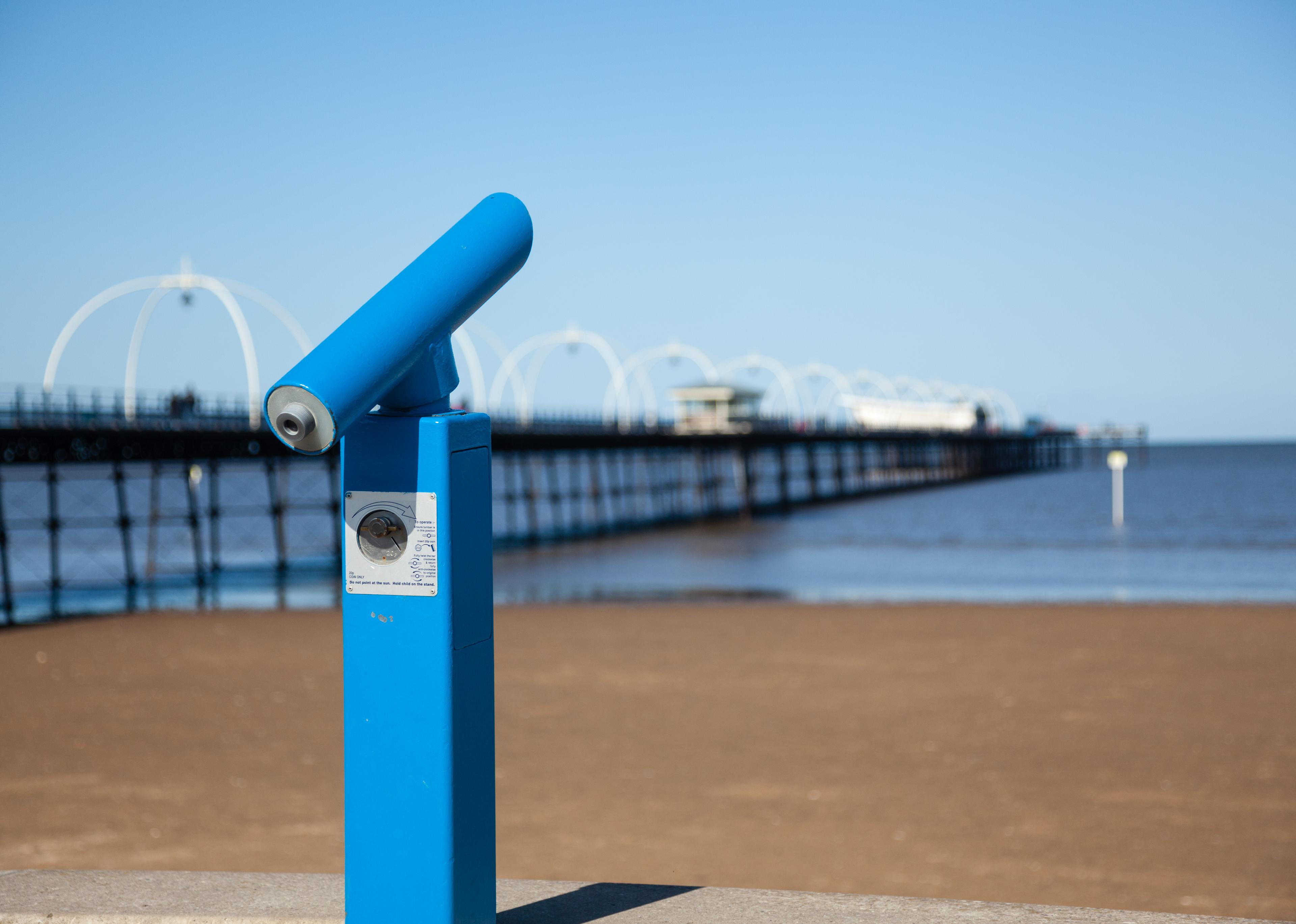 Blue binocular viewer facing a seaside pier against a clear blue sky.