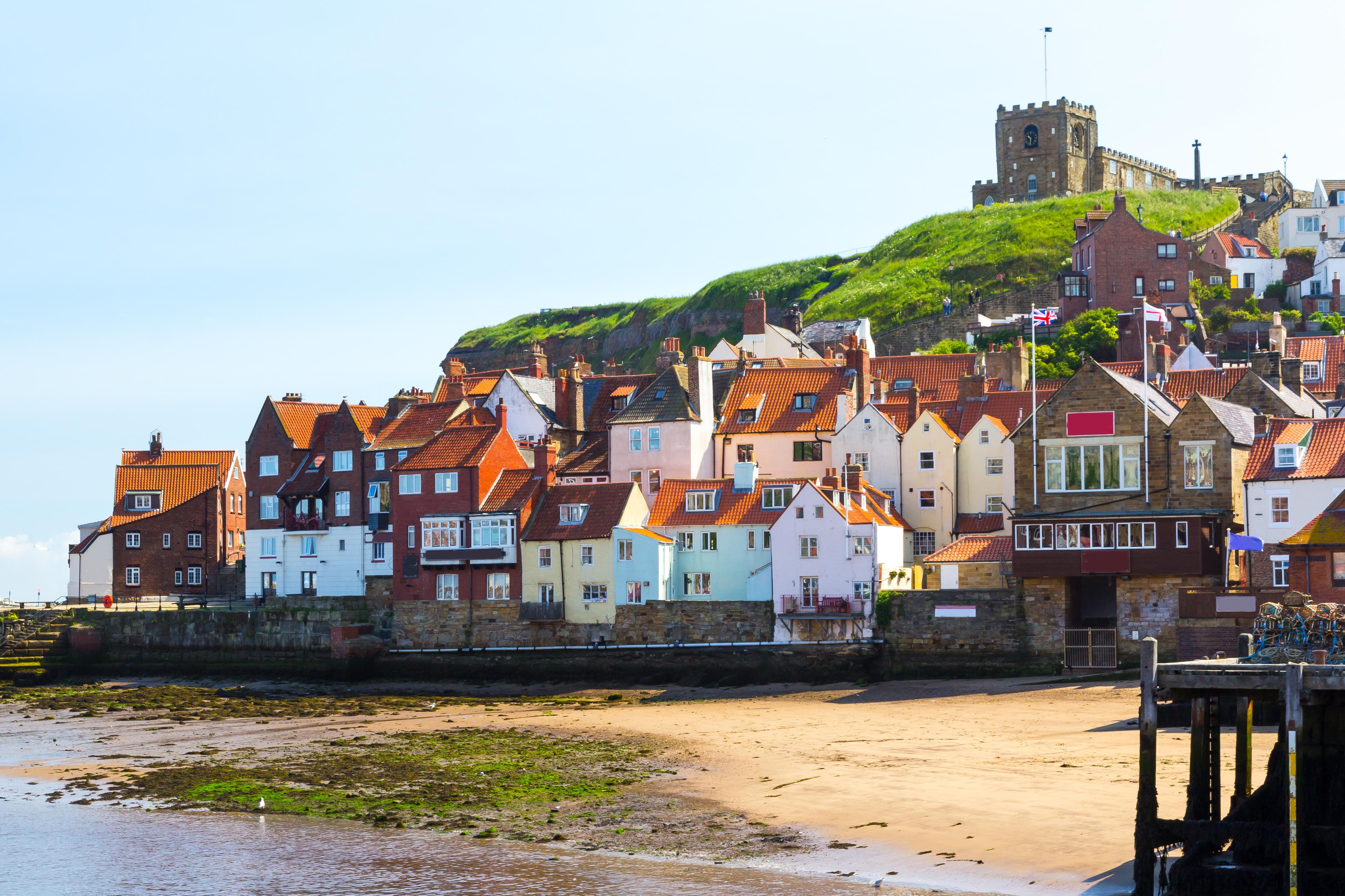 Coastal village with colorful houses on a hill, sandy shoreline, and a historic church overlooking from above.