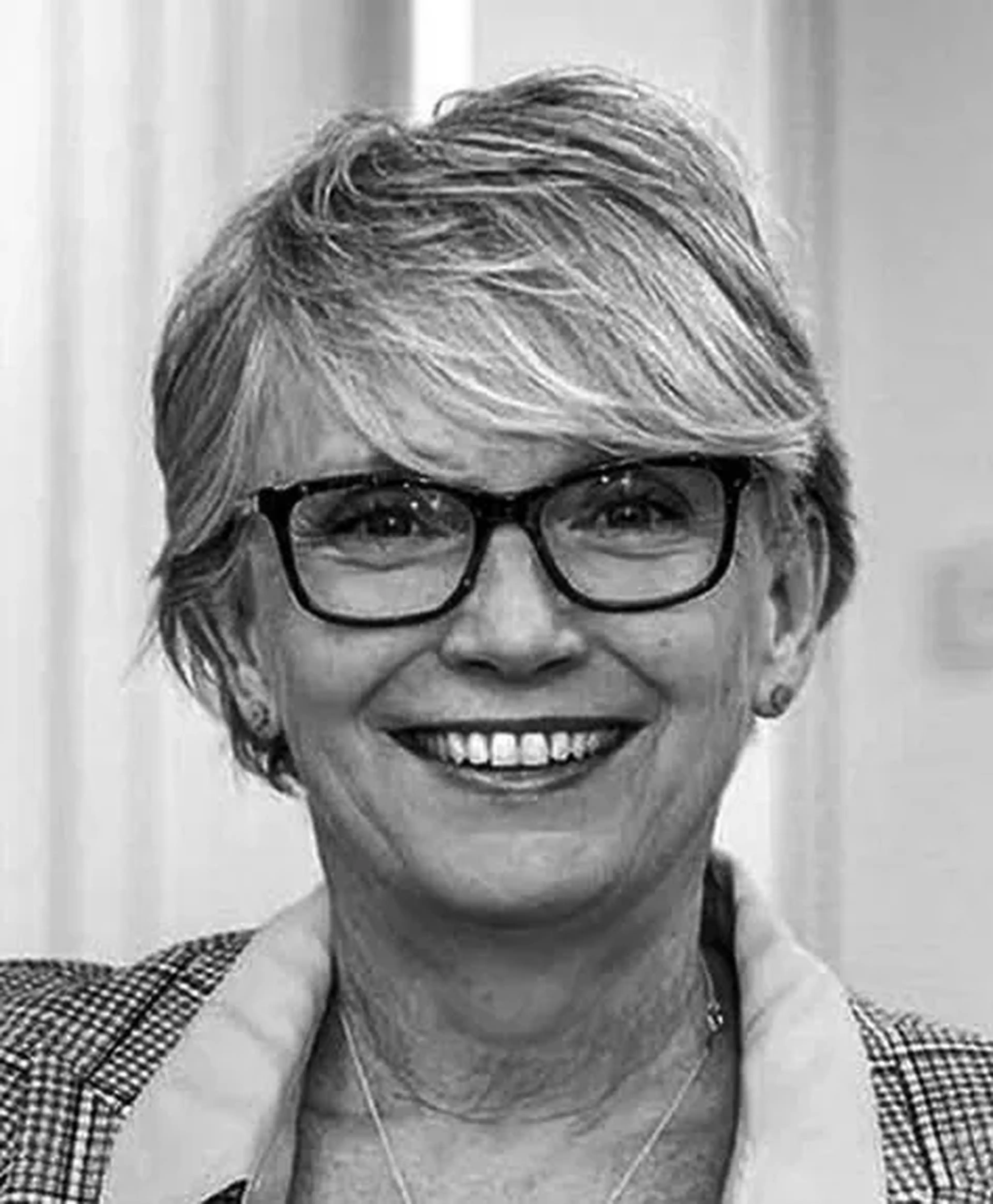 Smiling woman with short hair and glasses, wearing a plaid blazer and light shirt, in a black and white portrait.