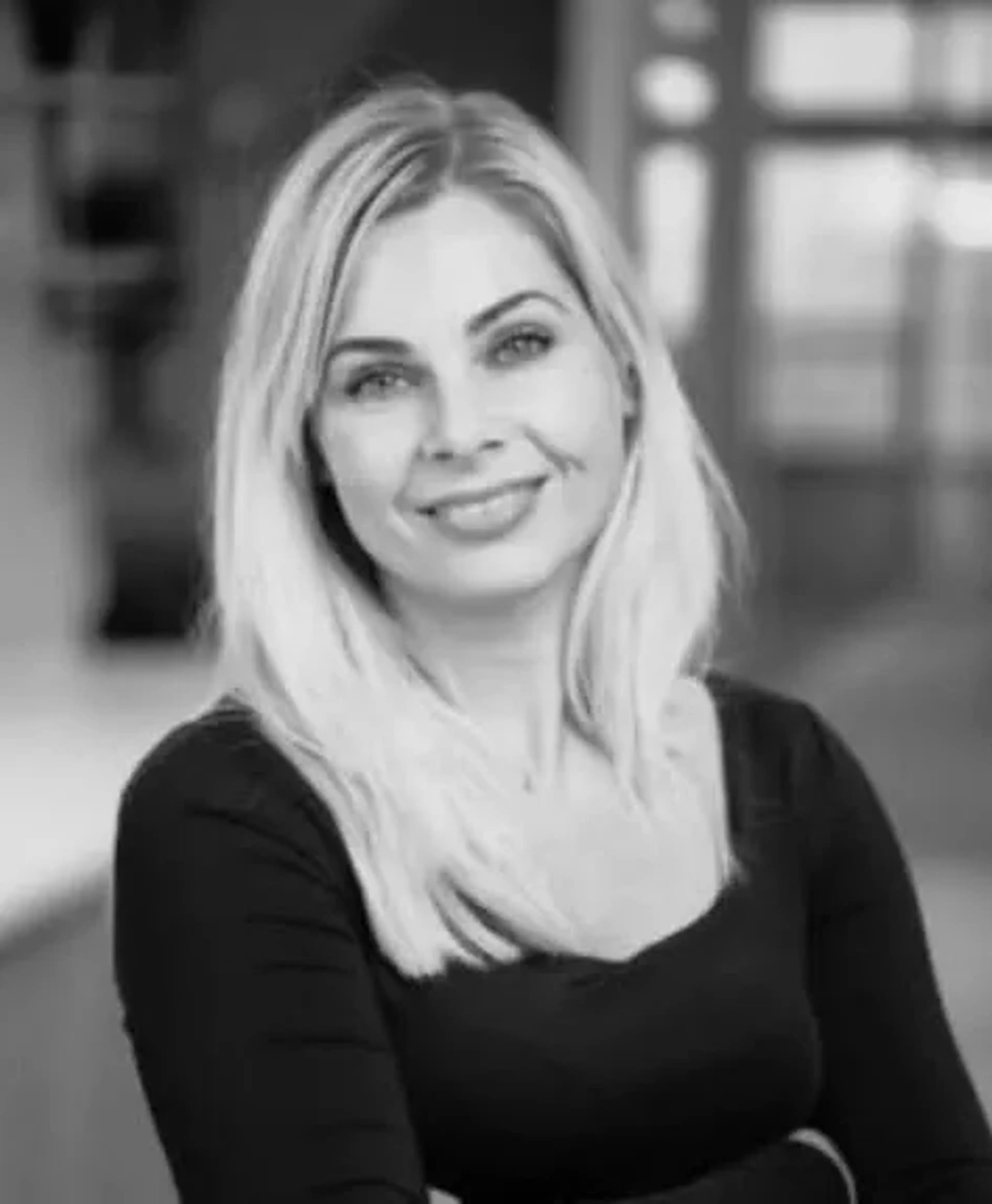 Smiling woman with long blonde hair, wearing a dark top, in a softly focused indoor setting.