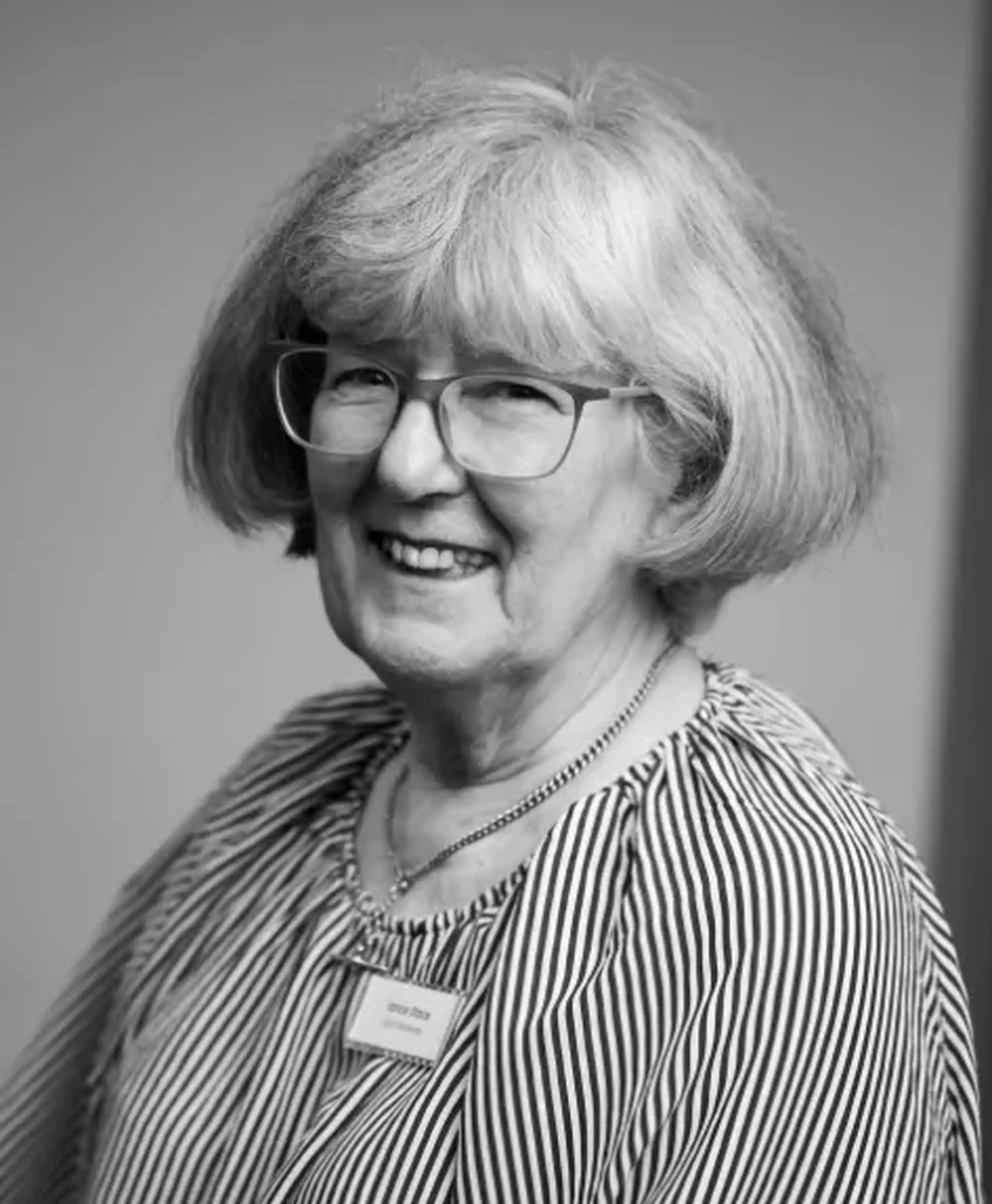 A smiling woman with glasses and short, gray hair, wearing a striped blouse and a name tag. The photo is in black and white.