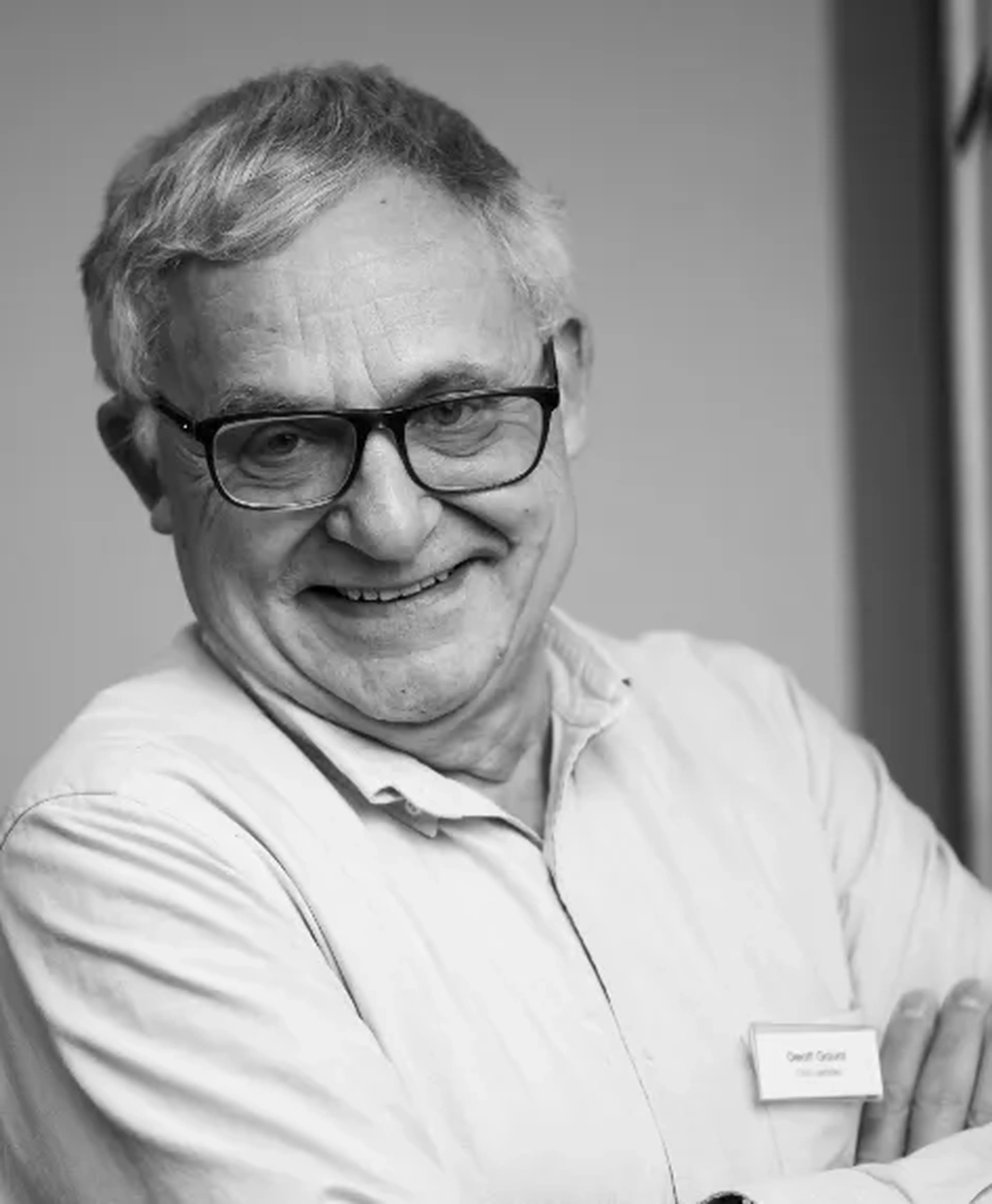 An older man smiling, wearing glasses and a light-colored shirt, with arms crossed. He's indoors, standing near a wall. Black and white image.