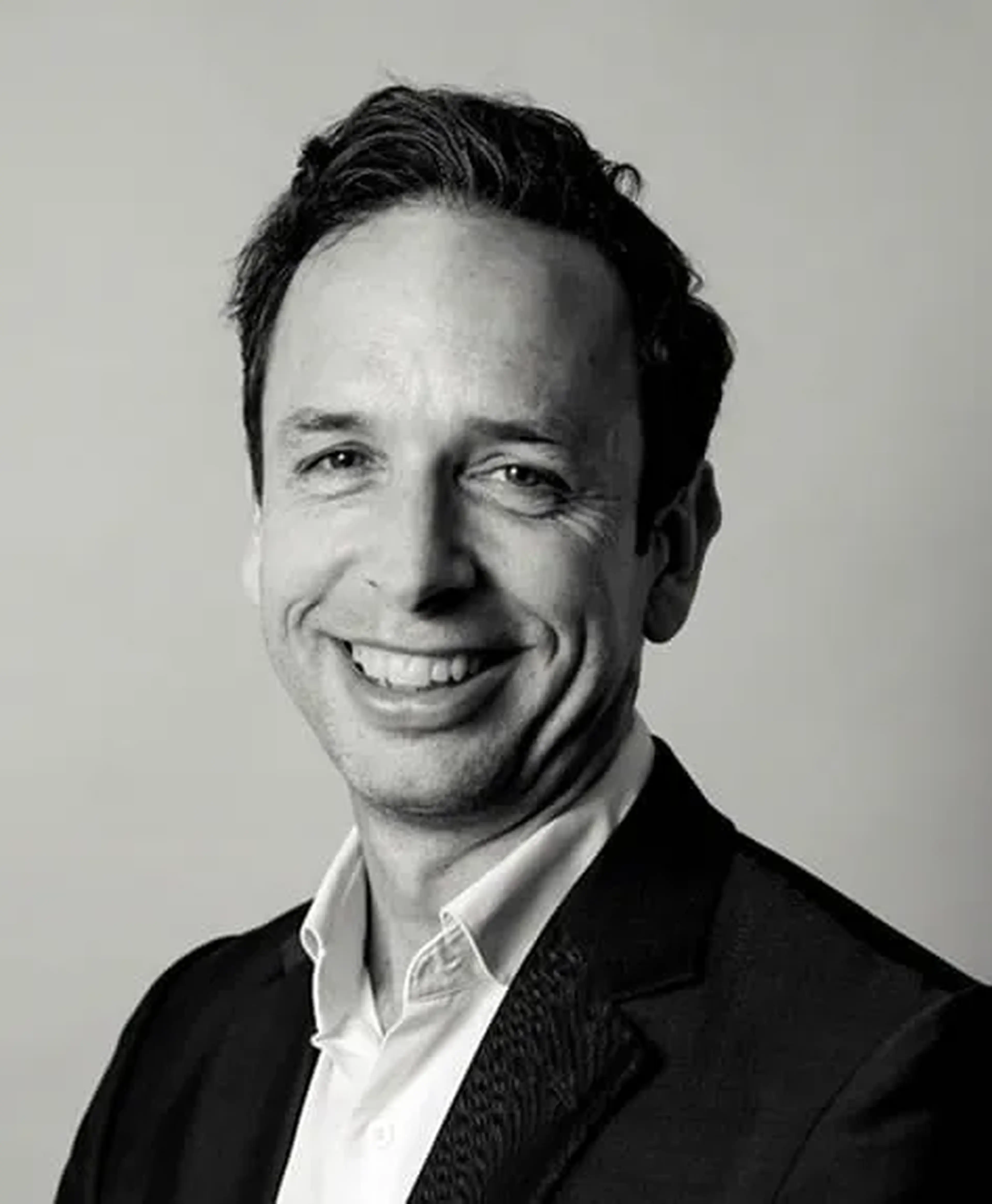 Smiling man in a suit with short hair against a plain background, photographed in black and white.