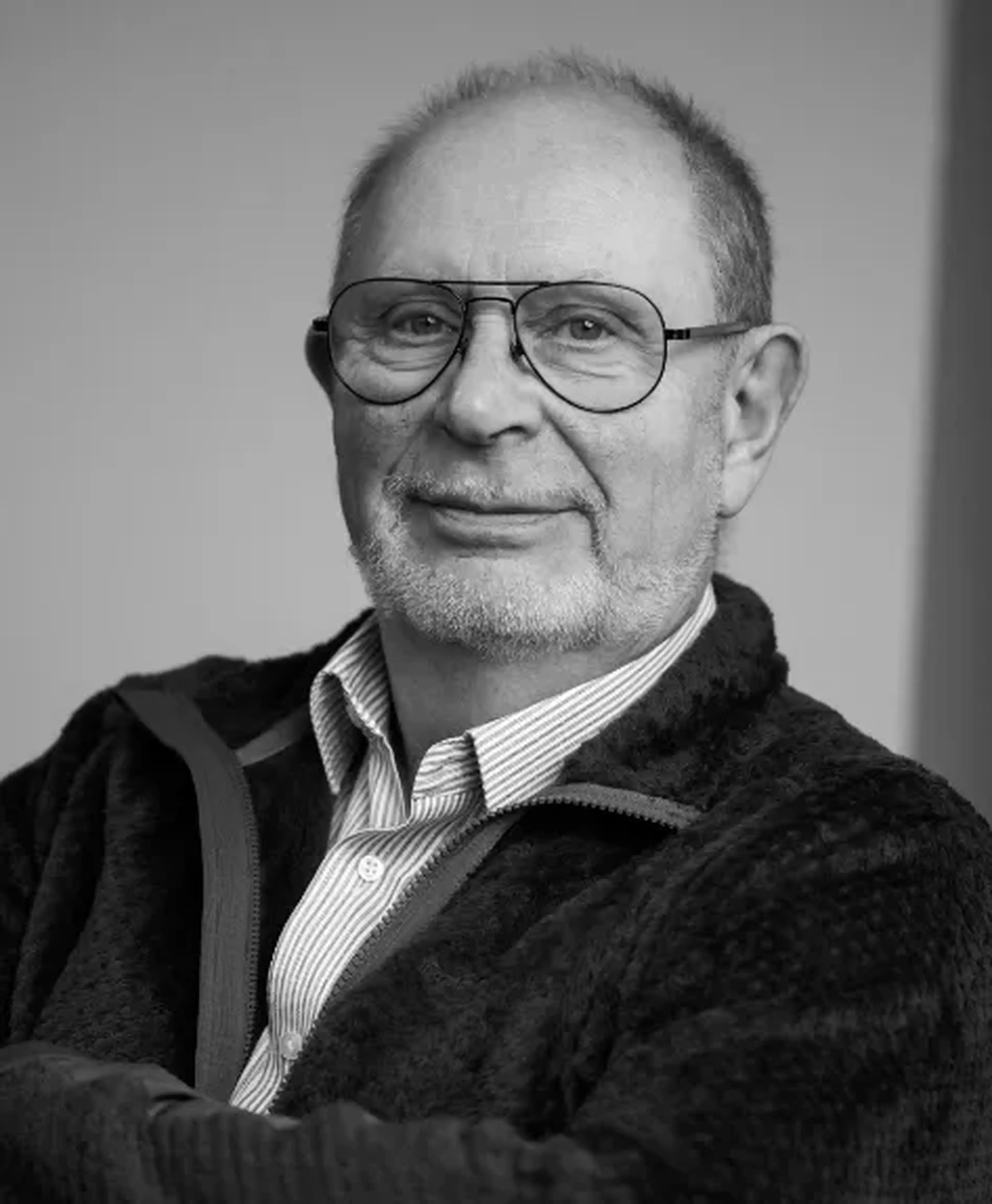 Black and white portrait of an older man with glasses, a beard, and a striped shirt under a zip-up jacket, smiling slightly.