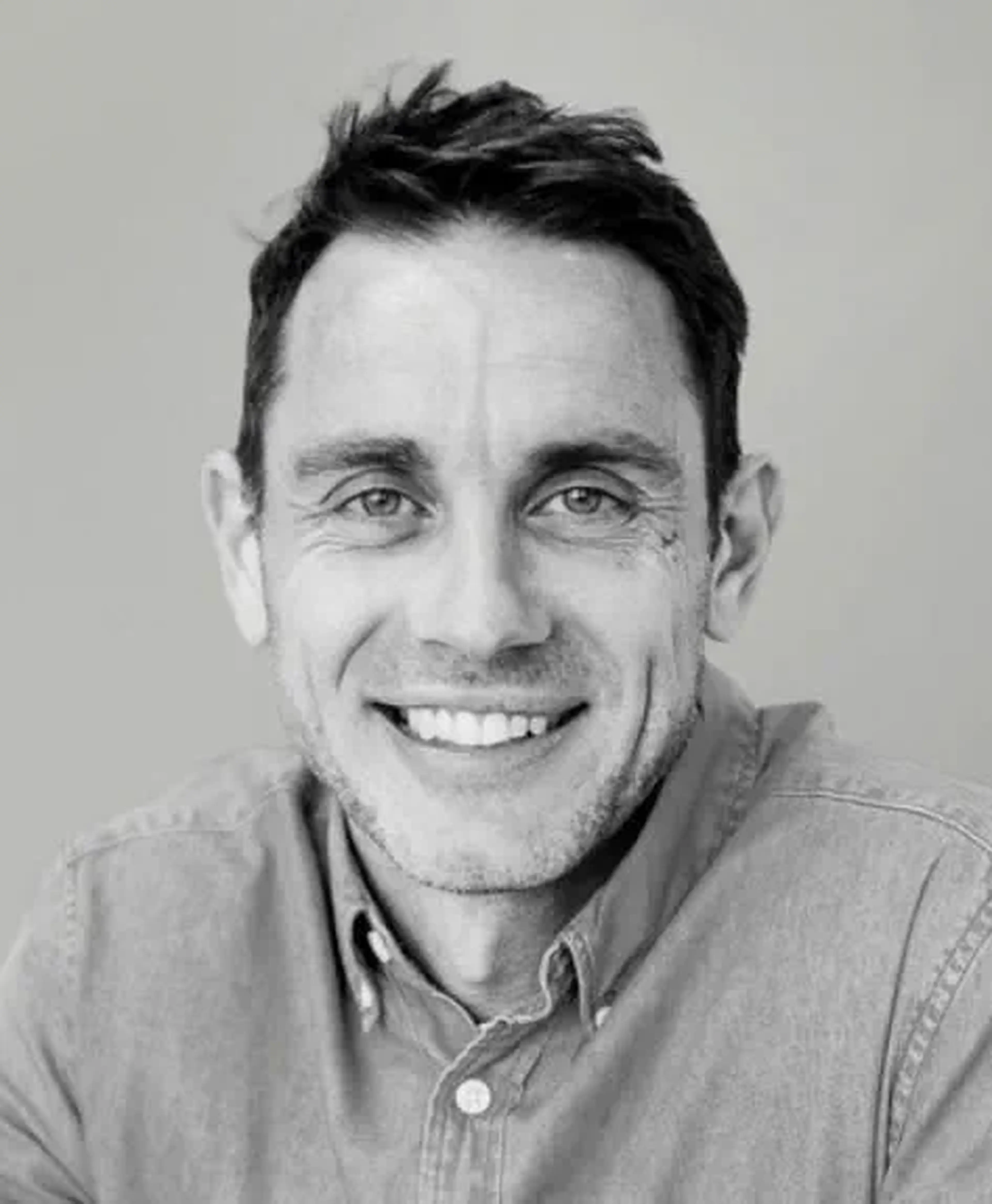 Smiling man with short hair wearing a denim shirt, looking directly at the camera against a neutral background.