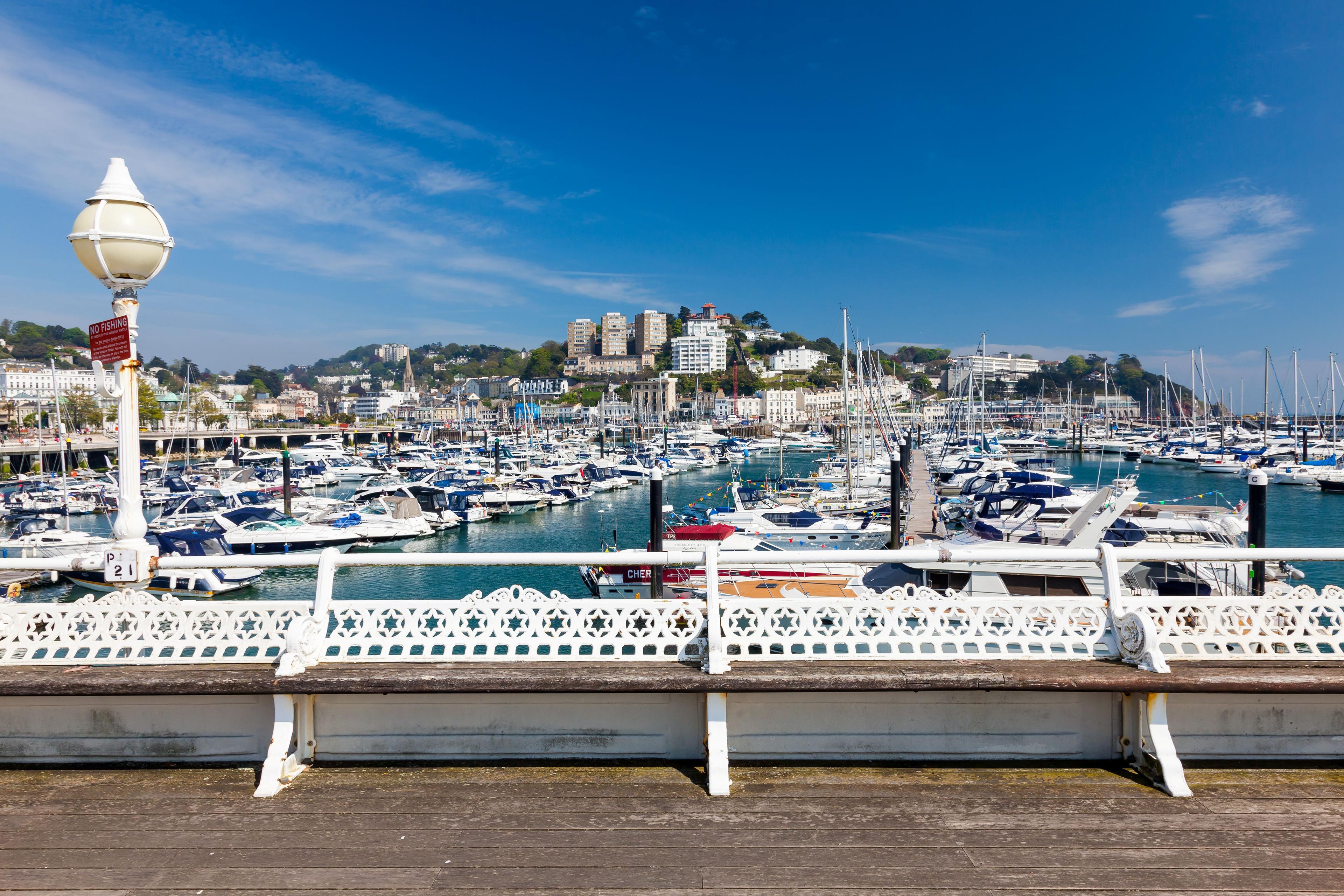 Bright seaside marina filled with sailboats and yachts, viewed over a decorative promenade railing toward a hillside town under blue skies.
