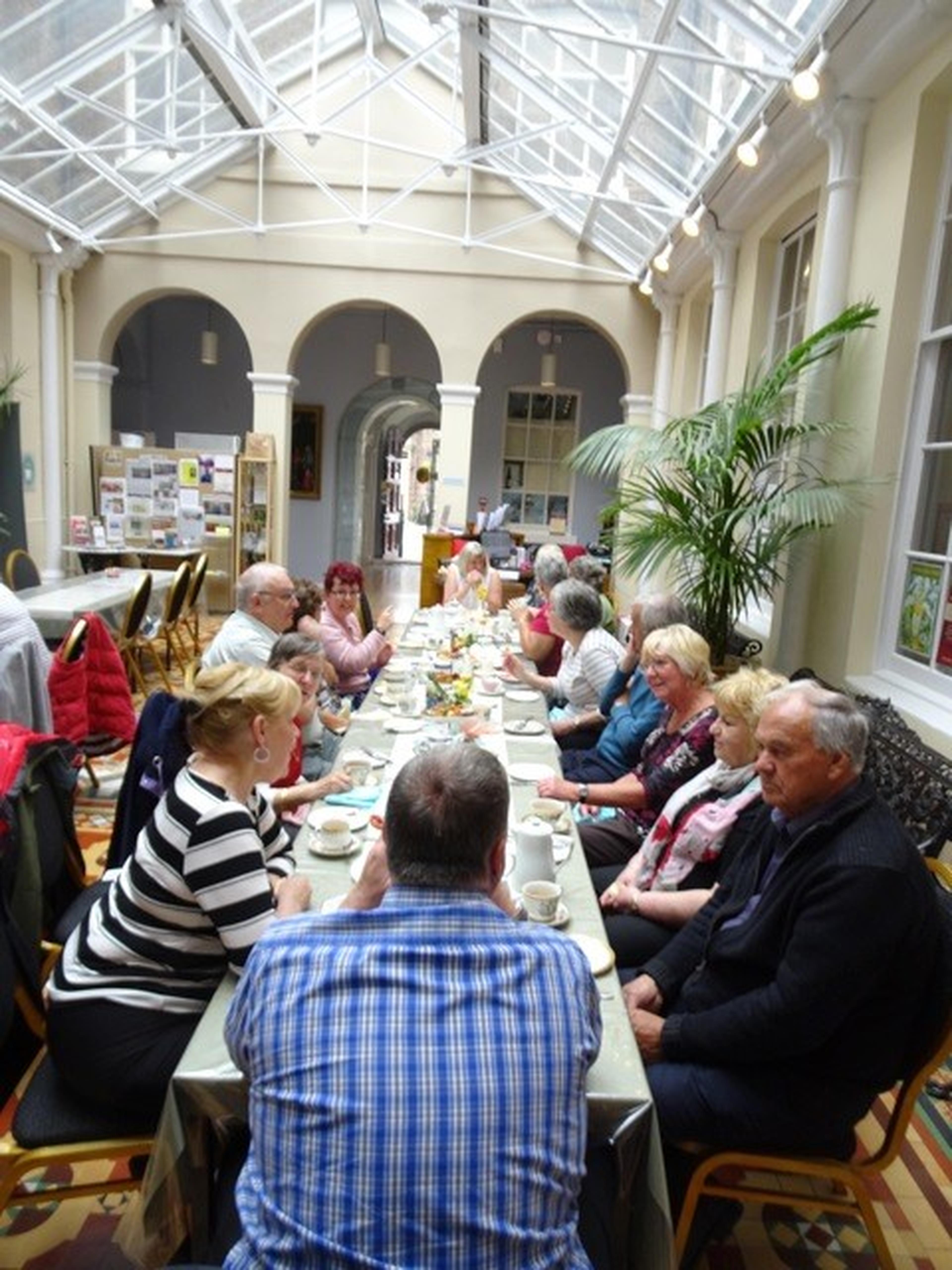 A group of people sitting around a long table inside a conservatory, having a meal and conversation