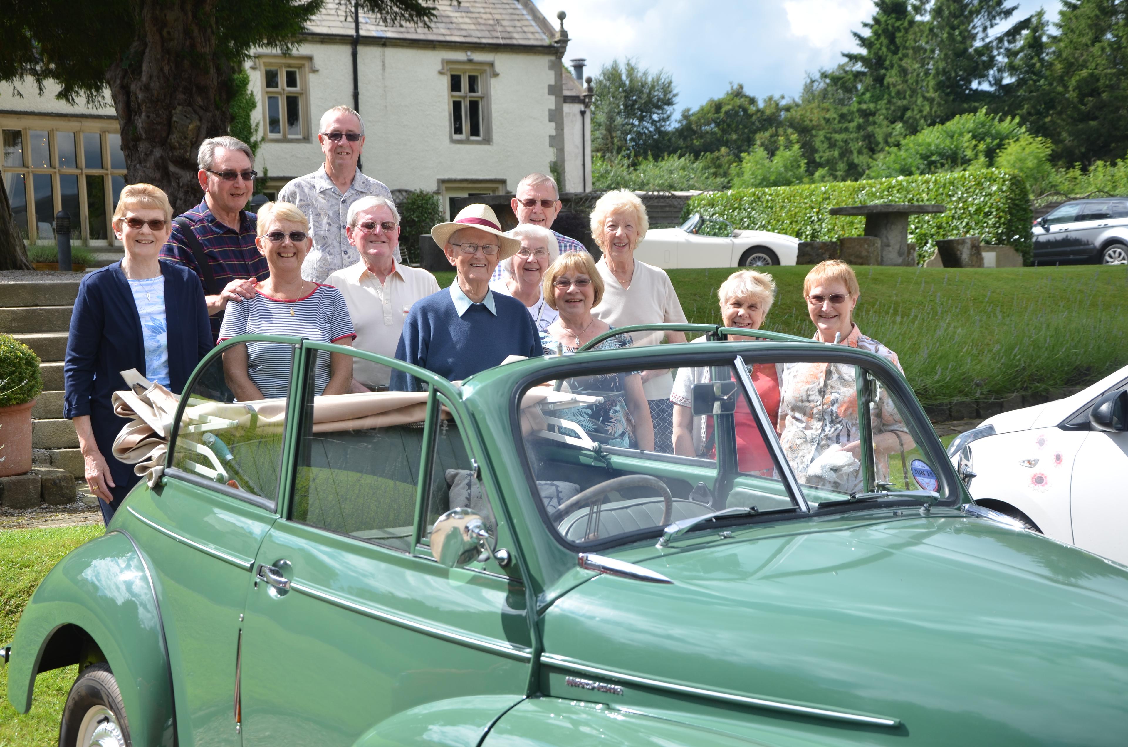 A group of smiling people stand together outdoors behind a classic car