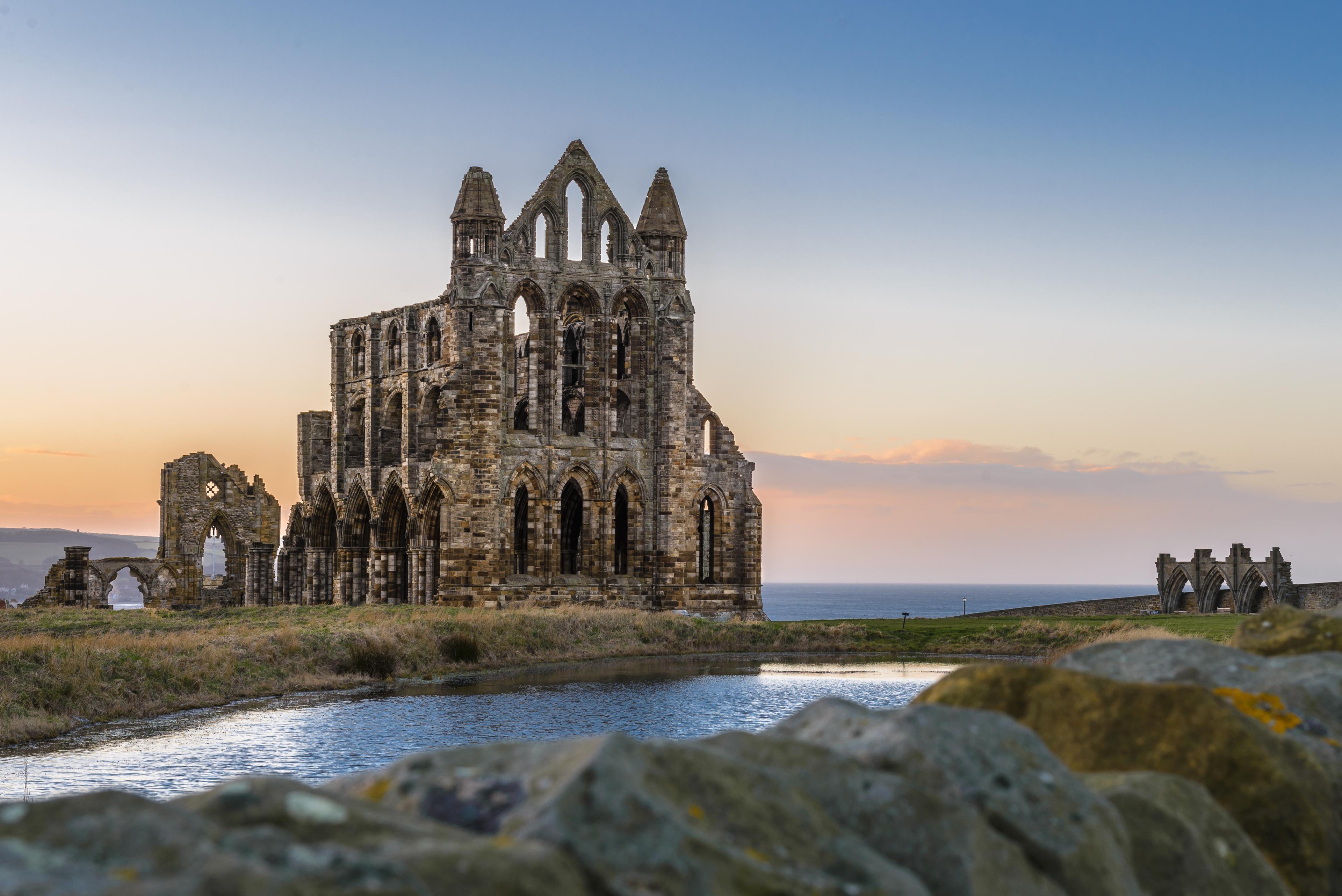 Ancient stone abbey ruins at sunset near a calm body of water, with a clear sky in the background.