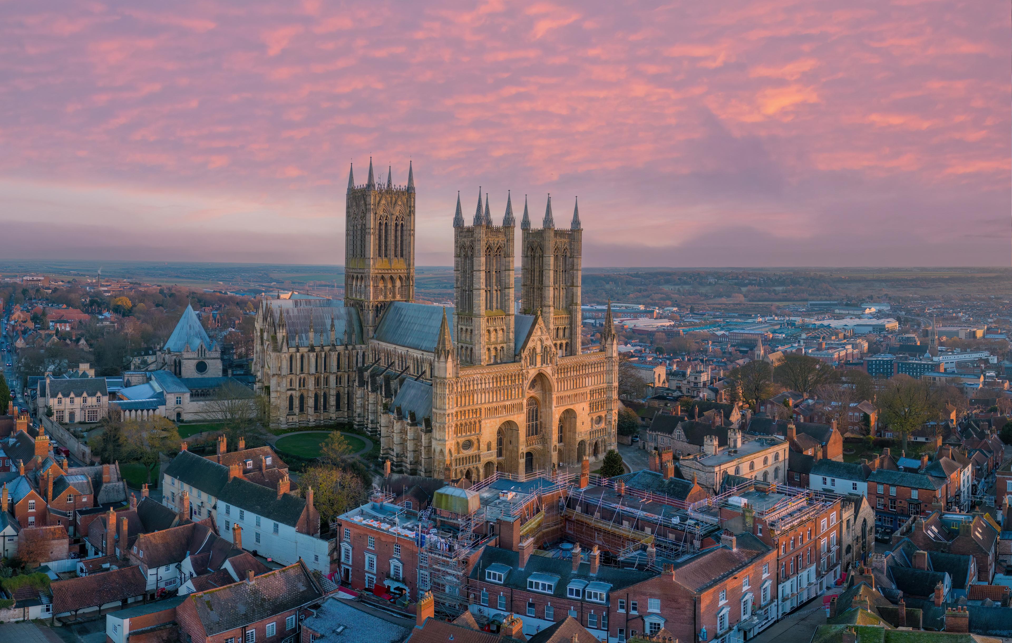Aerial view of a grand cathedral with multiple towers, surrounded by historic buildings under a vibrant pink and orange sunset sky.