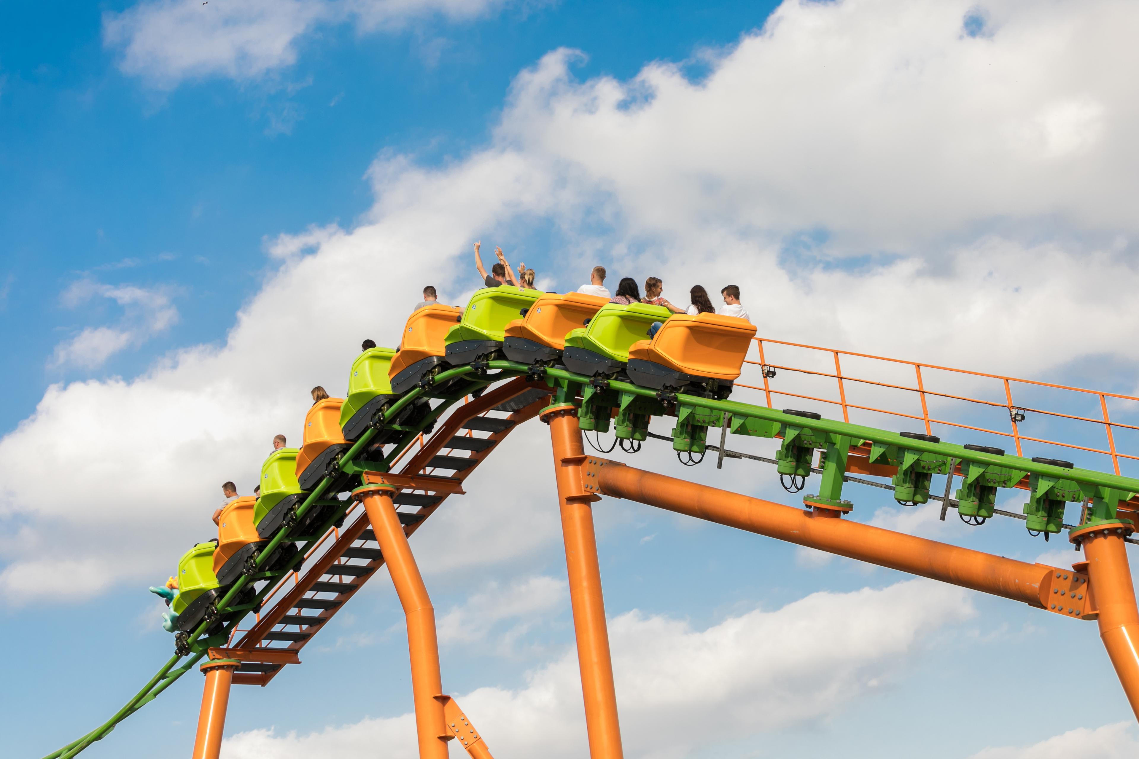 Bright green and orange roller coaster cars crest a steep track against a blue, cloudy sky, carrying riders with raised arms in a thrilling, joyful moment.