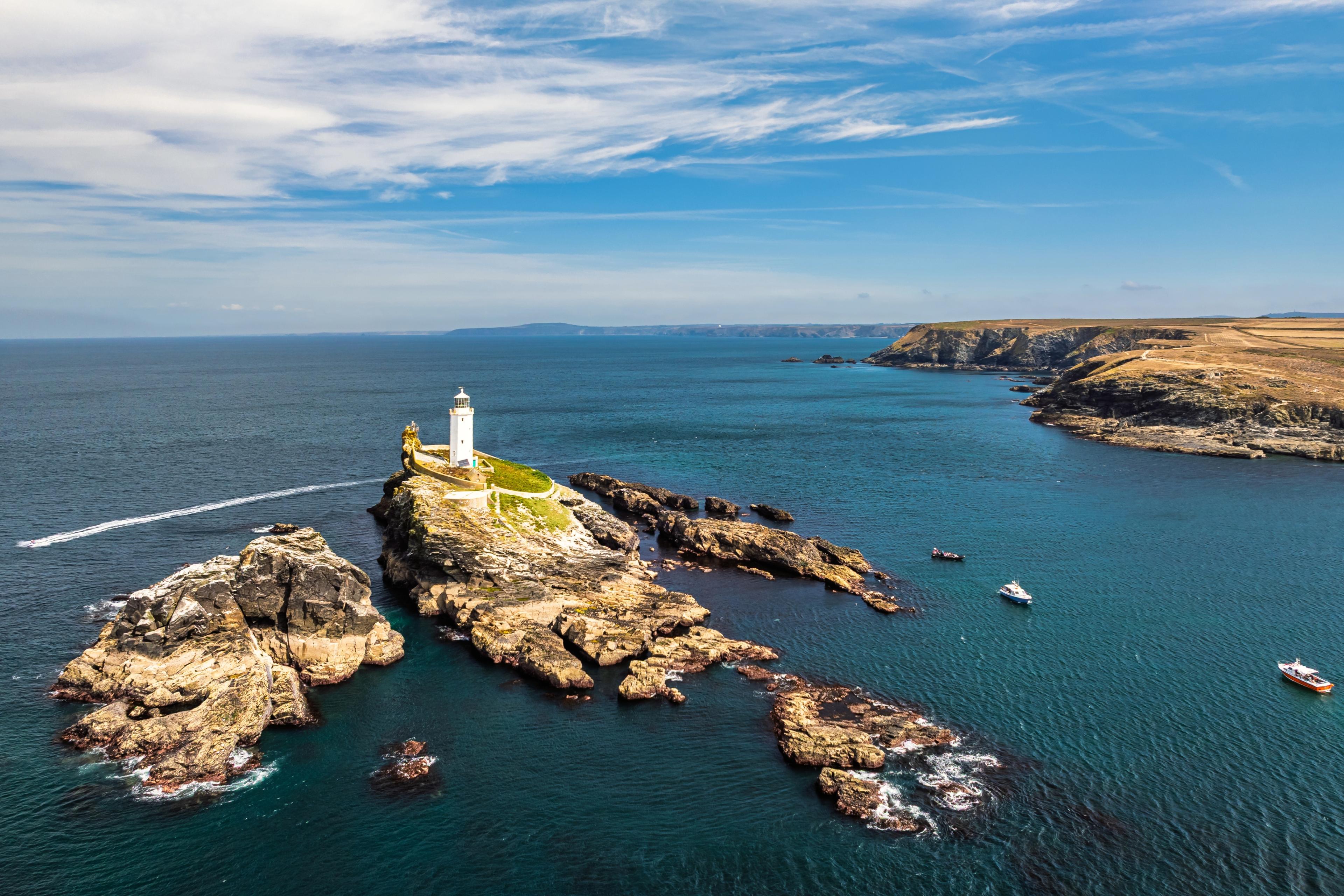 A coastal lighthouse on rocky cliffs surrounded by blue ocean, under a clear sky with boats nearby.