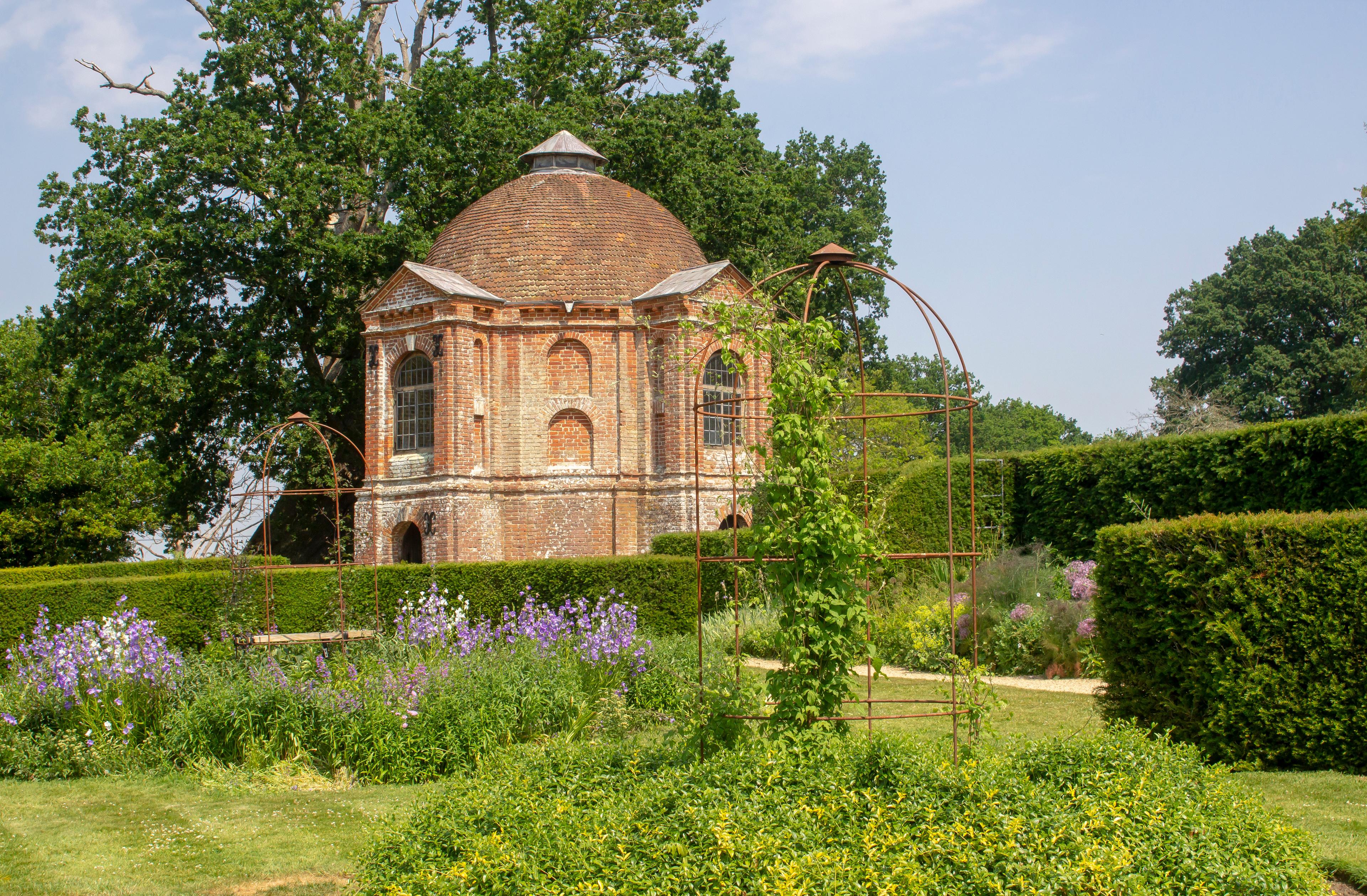 A brick dome building surrounded by lush greenery and flowering plants, set against a backdrop of trees under a clear sky.
