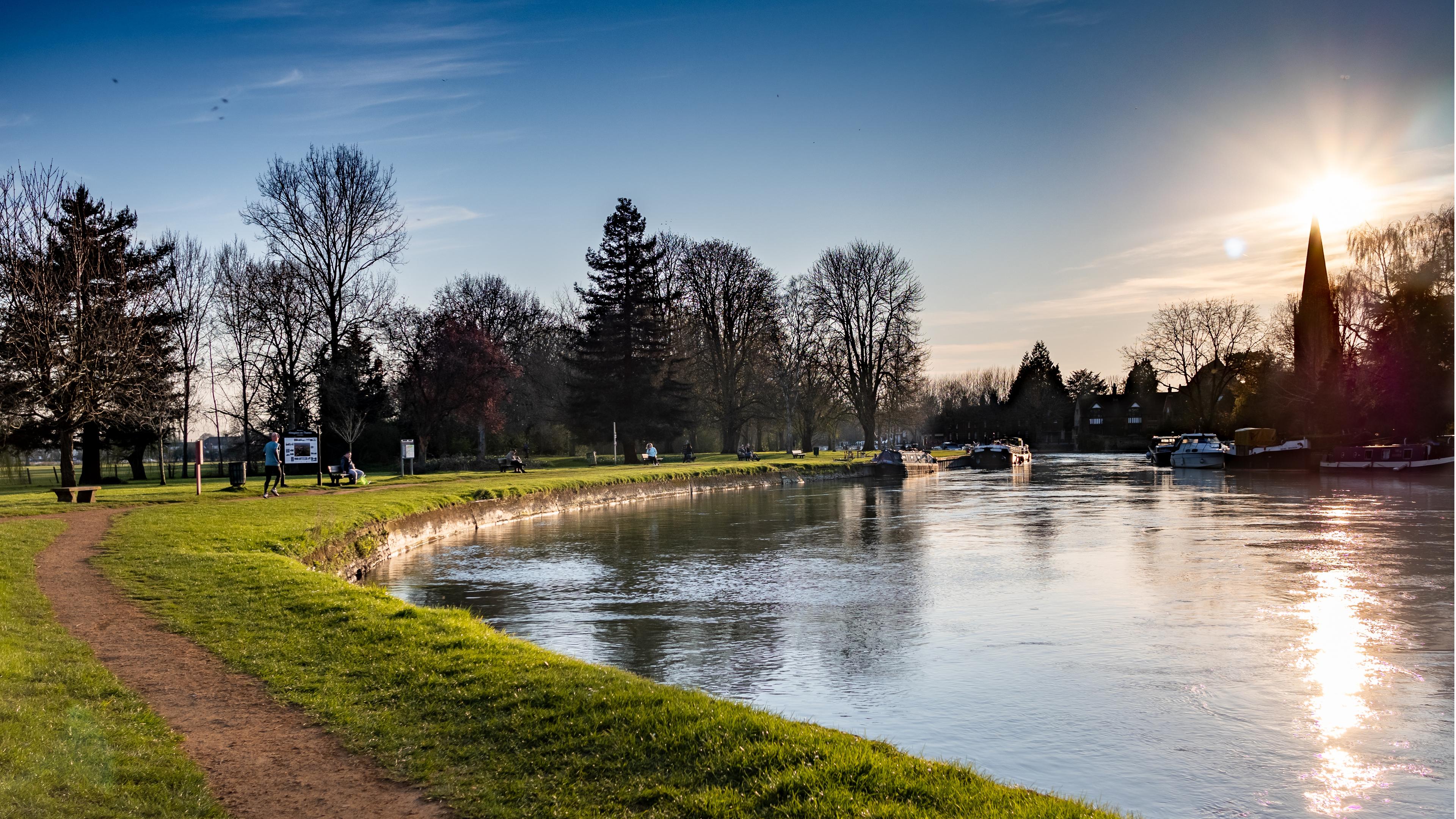 Riverside scene at sunset with trees and boats under a clear sky. A dirt path follows the riverbank, bordered by green grass.