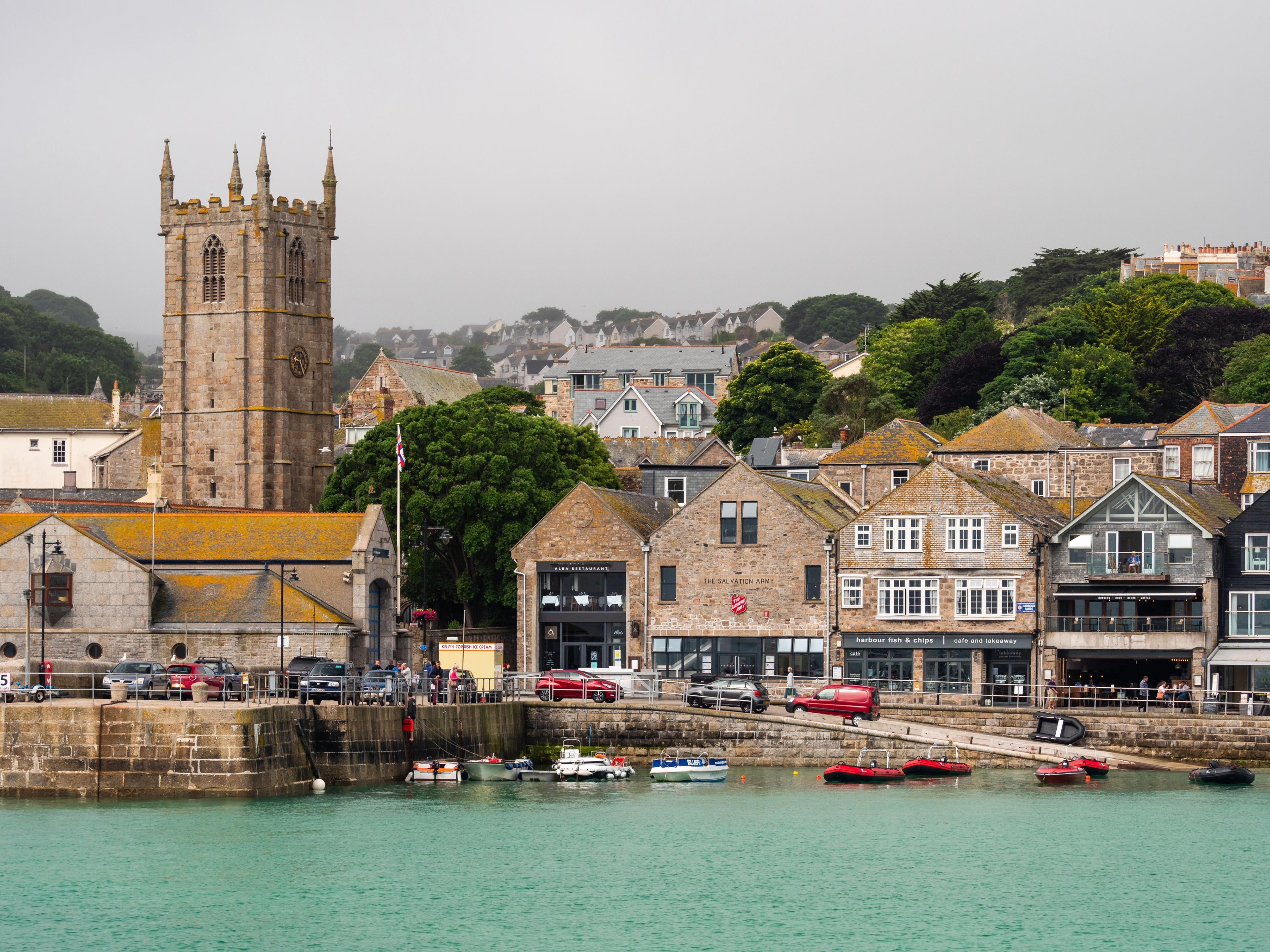 Coastal town with stone buildings and church tower, overlooking a marina with small boats on turquoise water and a hilly backdrop.