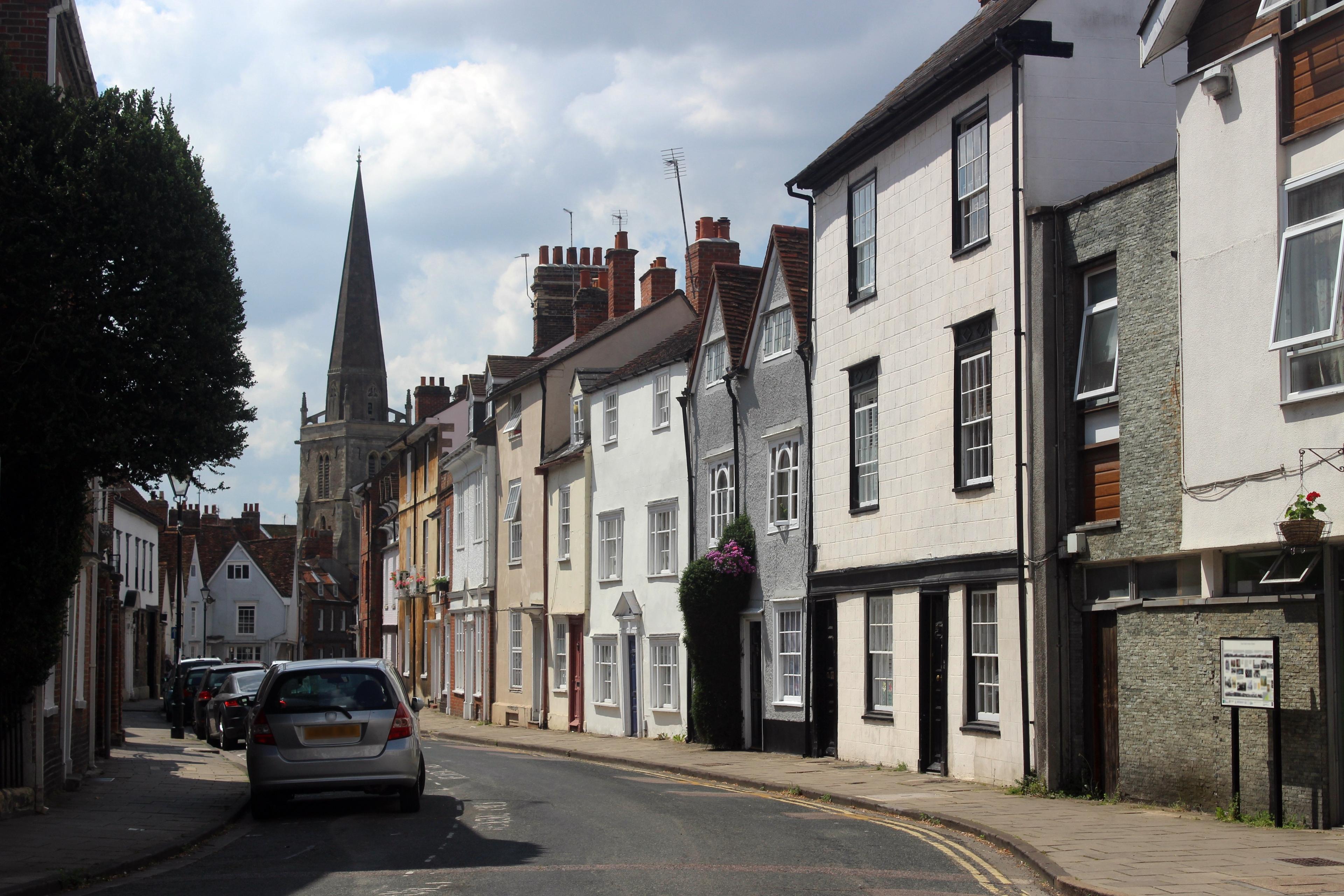 A quaint street with historic row houses, a parked car, and a church spire visible in the background under a partly cloudy sky.