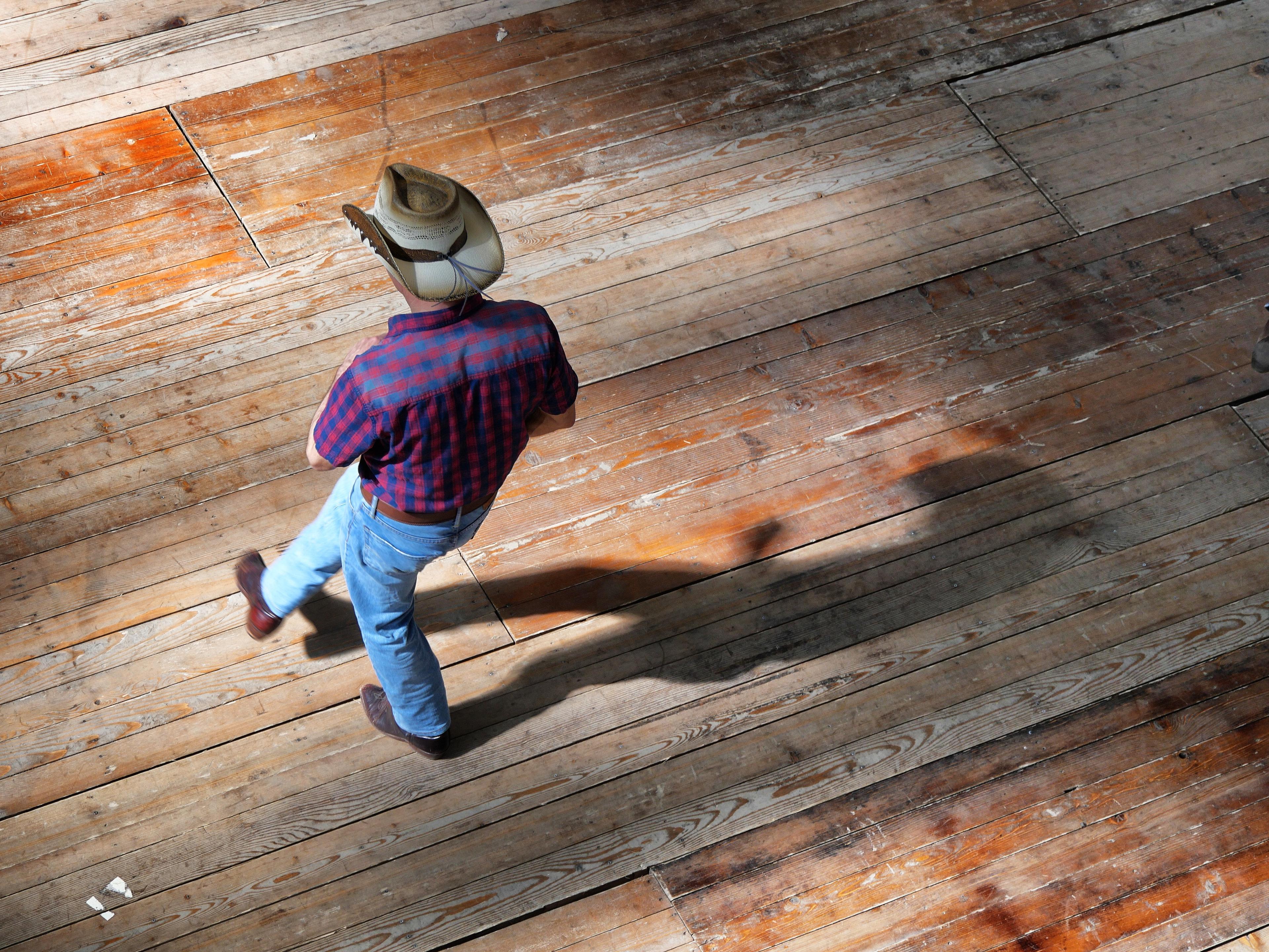 A person wearing a cowboy hat and plaid shirt dances solo on a sunlit wooden floor, casting a long shadow.