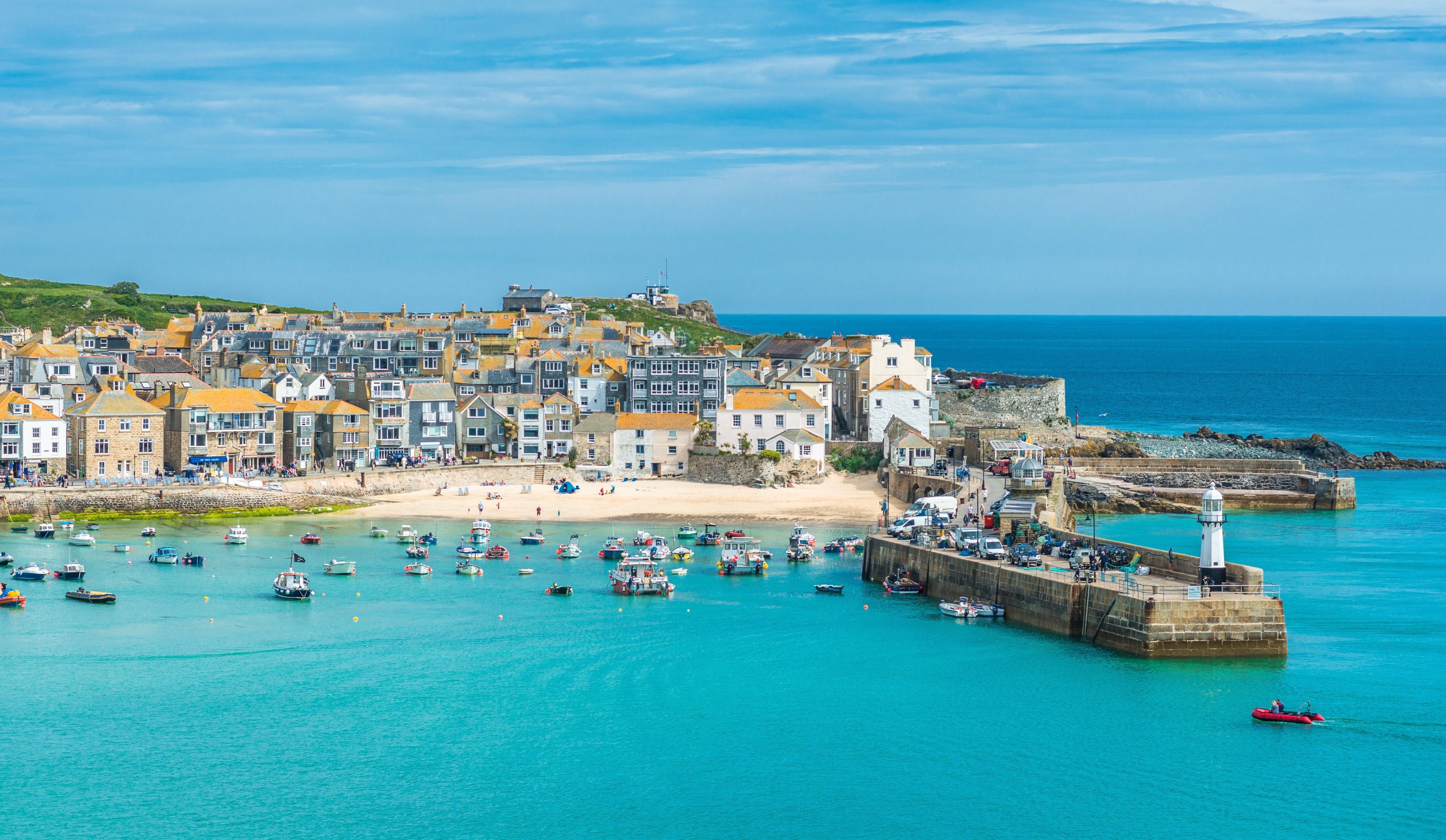 Coastal town with colorful buildings, a sandy beach, a harbor filled with boats, and a lighthouse, all under a blue sky.