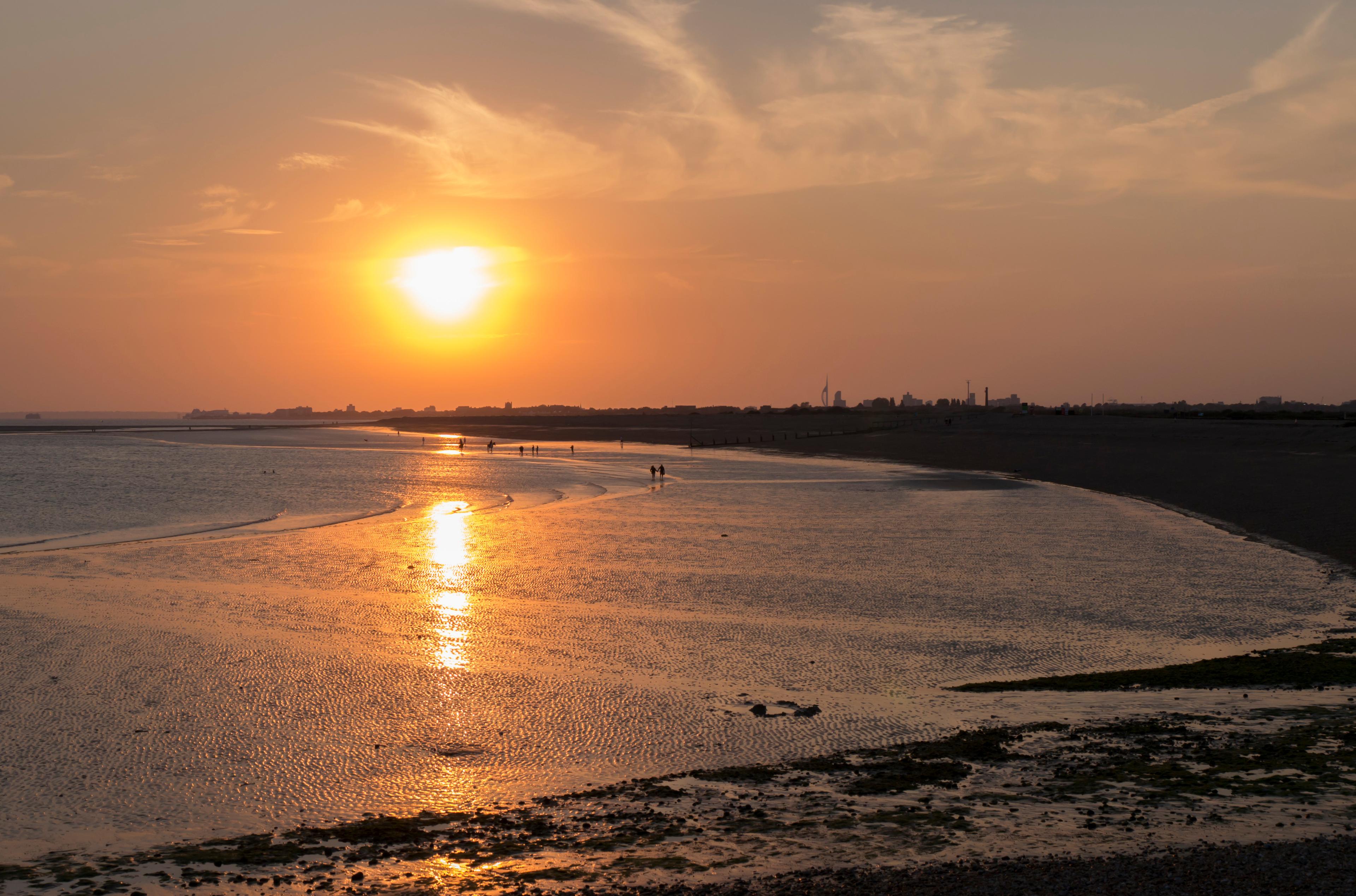 Golden sunset over a calm bay, casting a glowing reflection across wet sand, with distant shoreline silhouettes and a peaceful atmosphere.