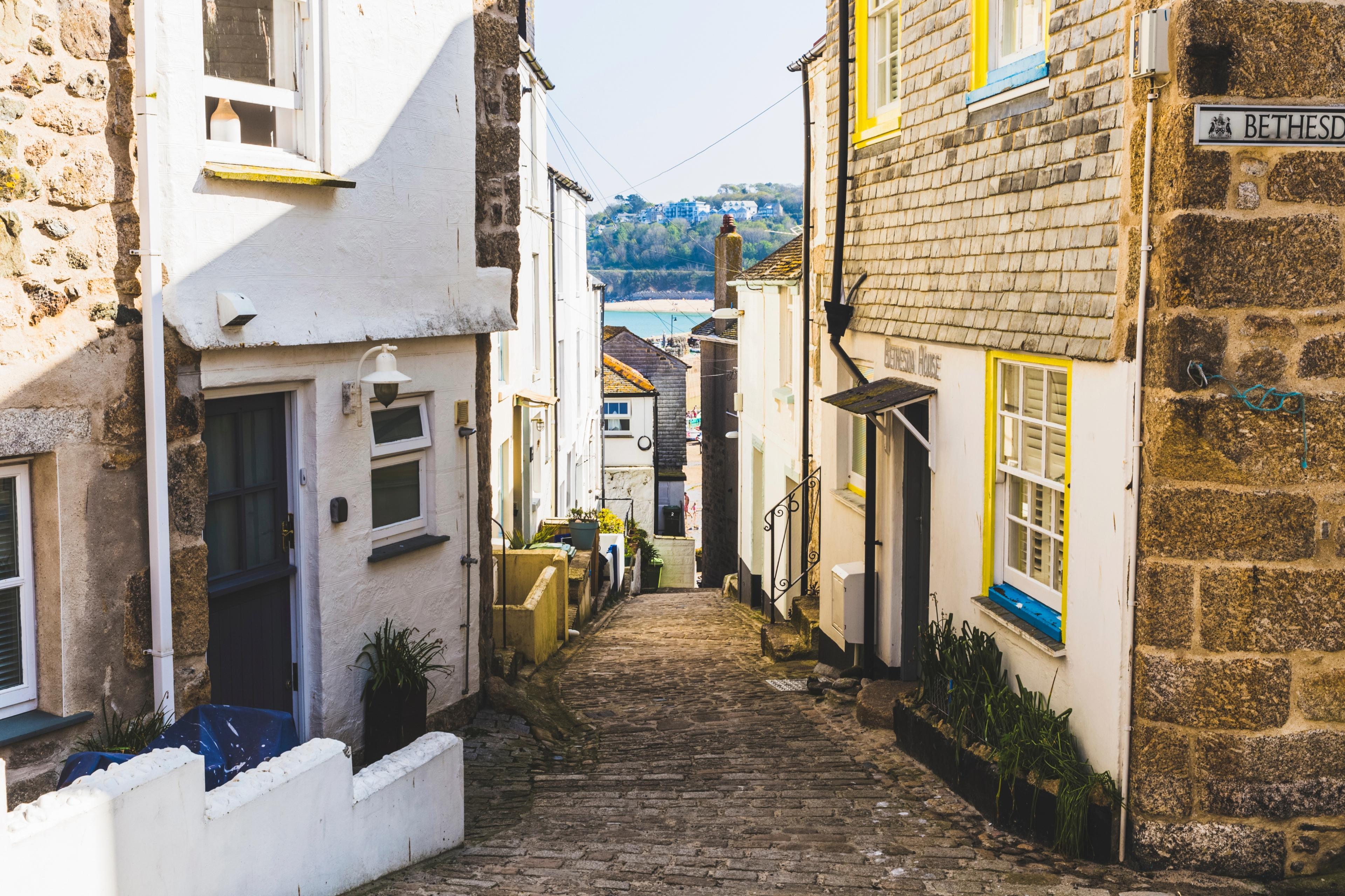 Narrow cobblestone alley with quaint houses, leading to a distant view of a serene bay under a clear blue sky.
