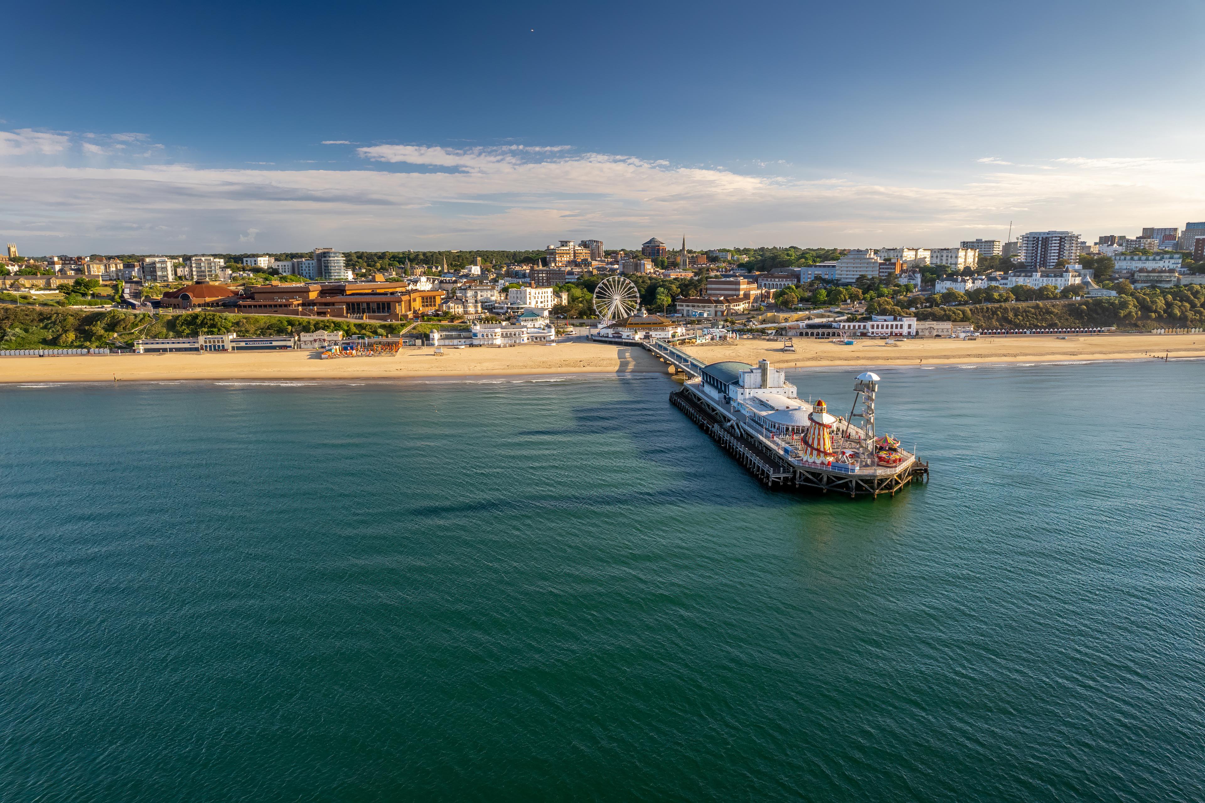 Aerial view of a coastal city with a pier extending into the calm sea under a clear blue sky, buildings and a Ferris wheel on the shoreline.