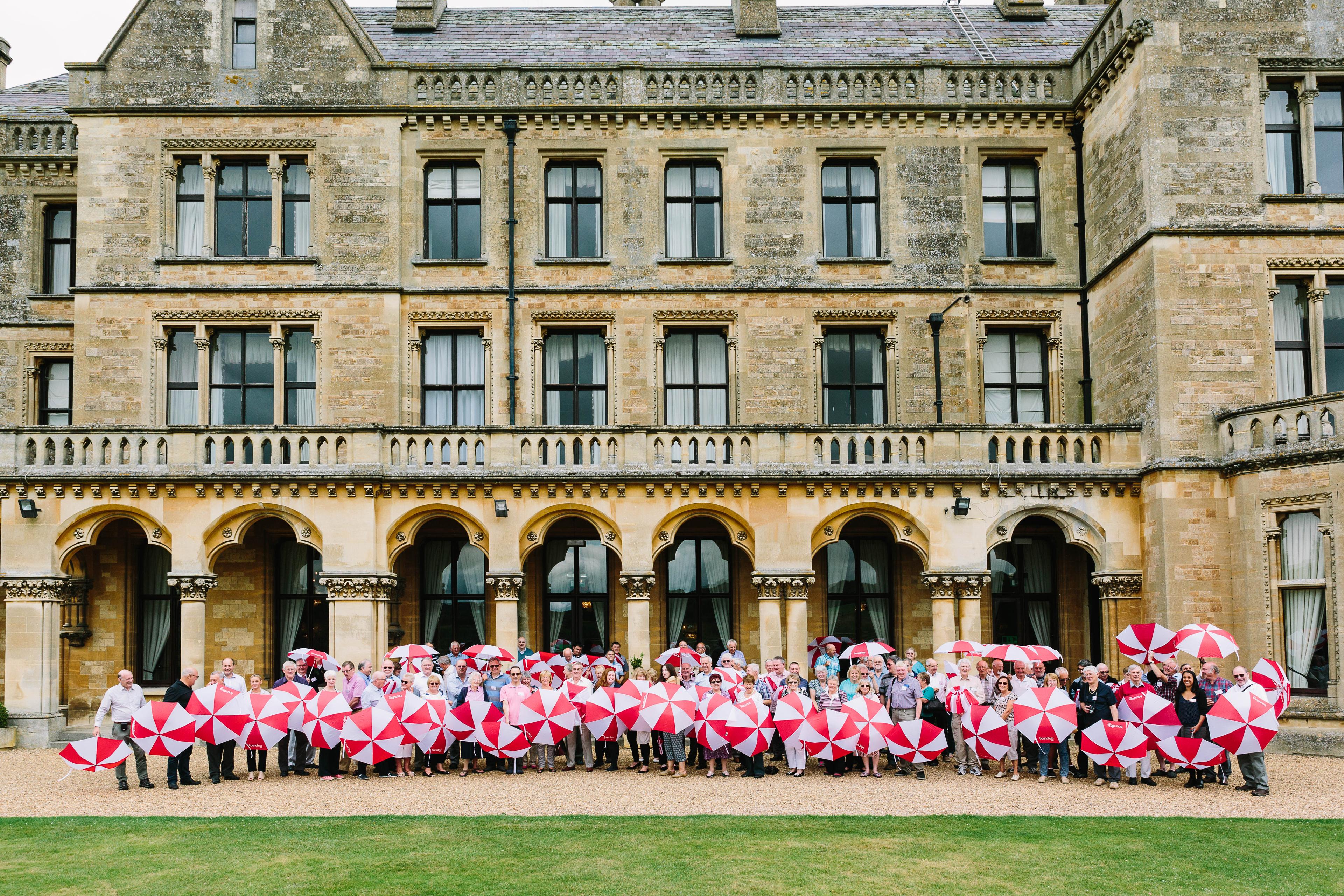 A large group of people holding red and white umbrellas stand outside a historic building with arched windows and stone facade.