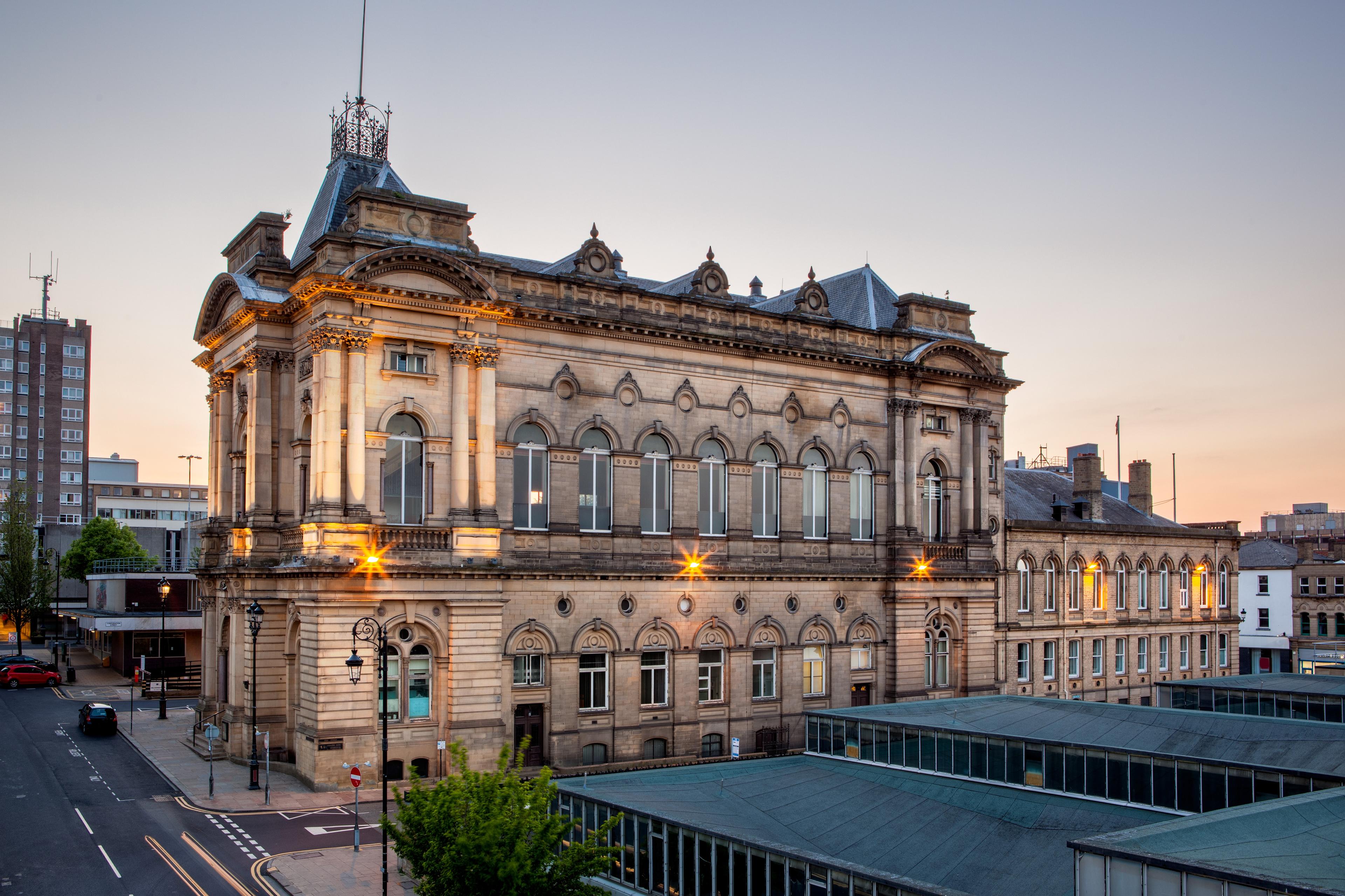 An ornate historic concert hall in central Huddersfield, West Yorkshire, with a grand stone façade, arched windows, decorative columns, and a distinctive rooftop tower, illuminated by warm evening lights above surrounding city streets.