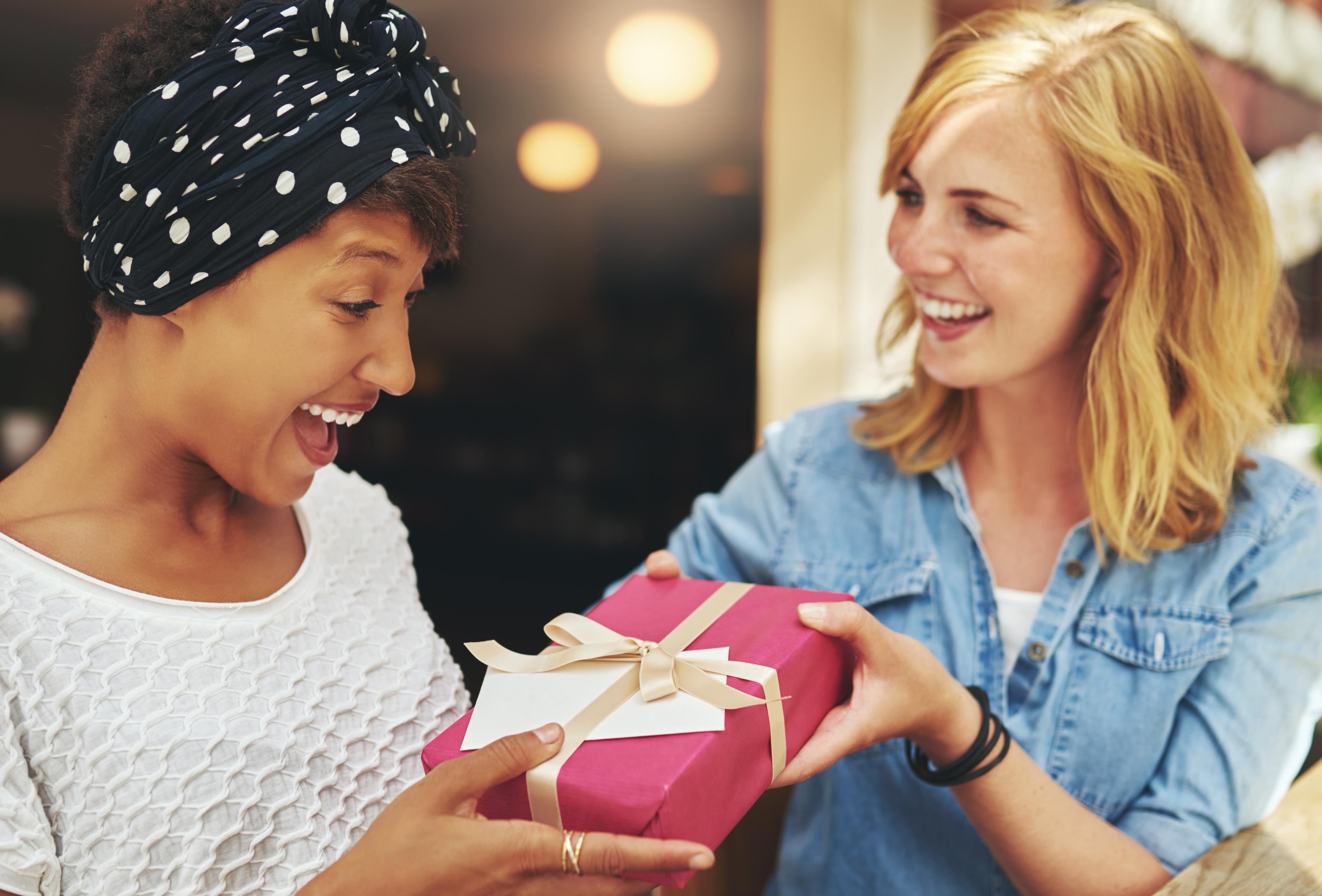 Two women smiling as one gives the other a gift wrapped in pink paper with a ribbon.