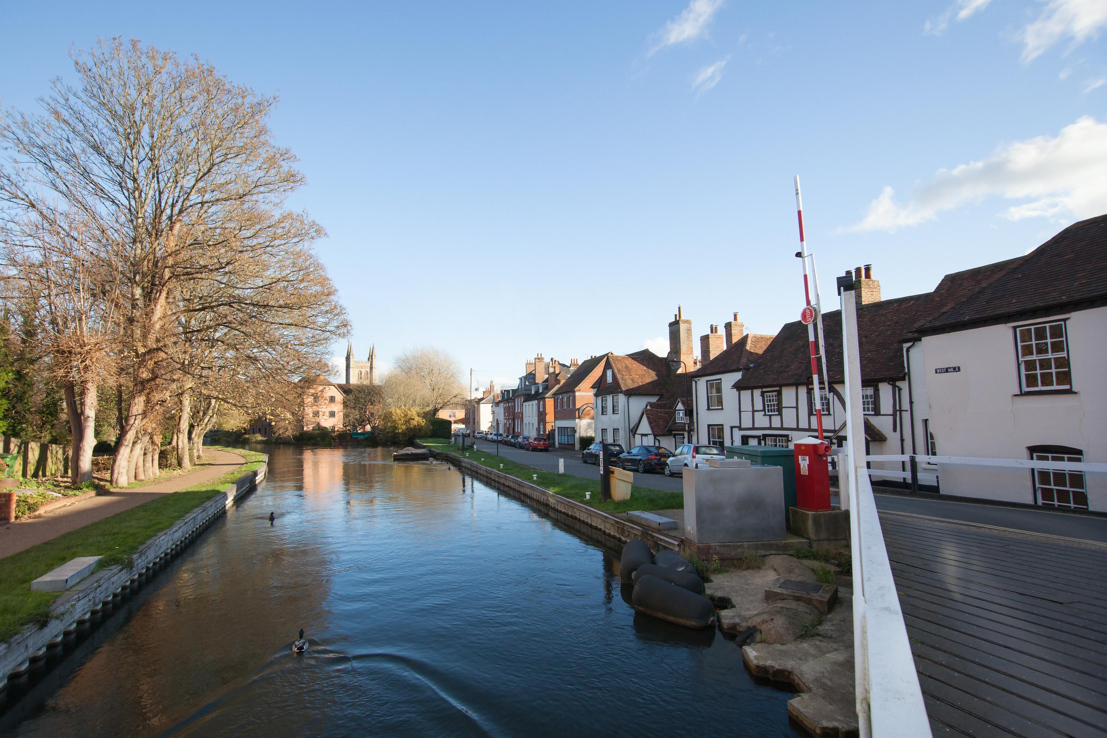 A scenic canal lined with charming historic houses on one side and trees on the other, under a clear blue sky.