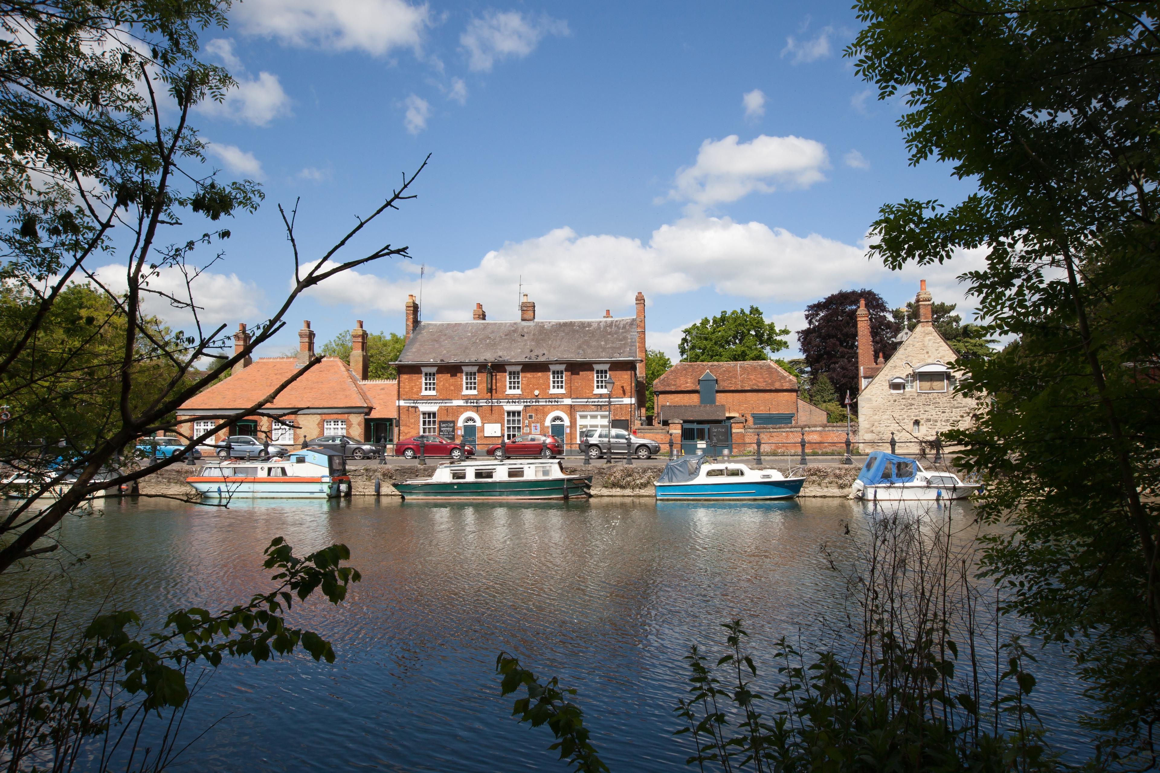 Riverside scene with moored boats, red-brick buildings, and a stone structure under a blue sky, framed by trees in the foreground.