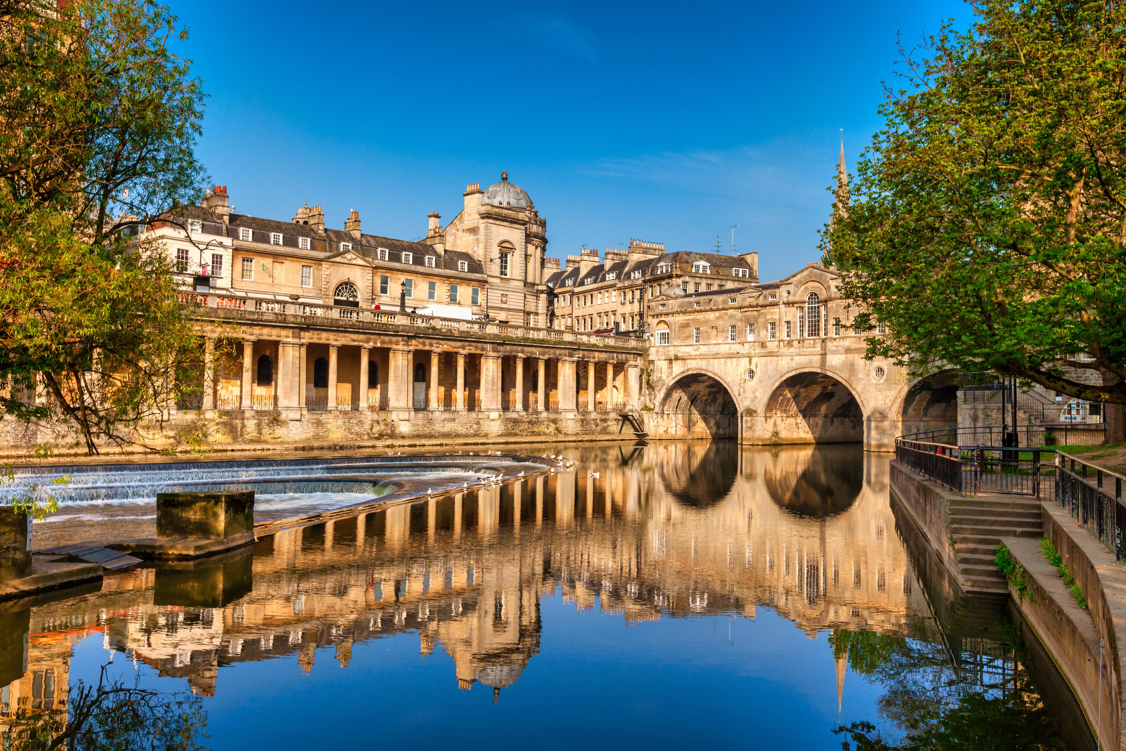 A historic stone bridge and buildings are reflected in a calm river, surrounded by trees under a clear blue sky.