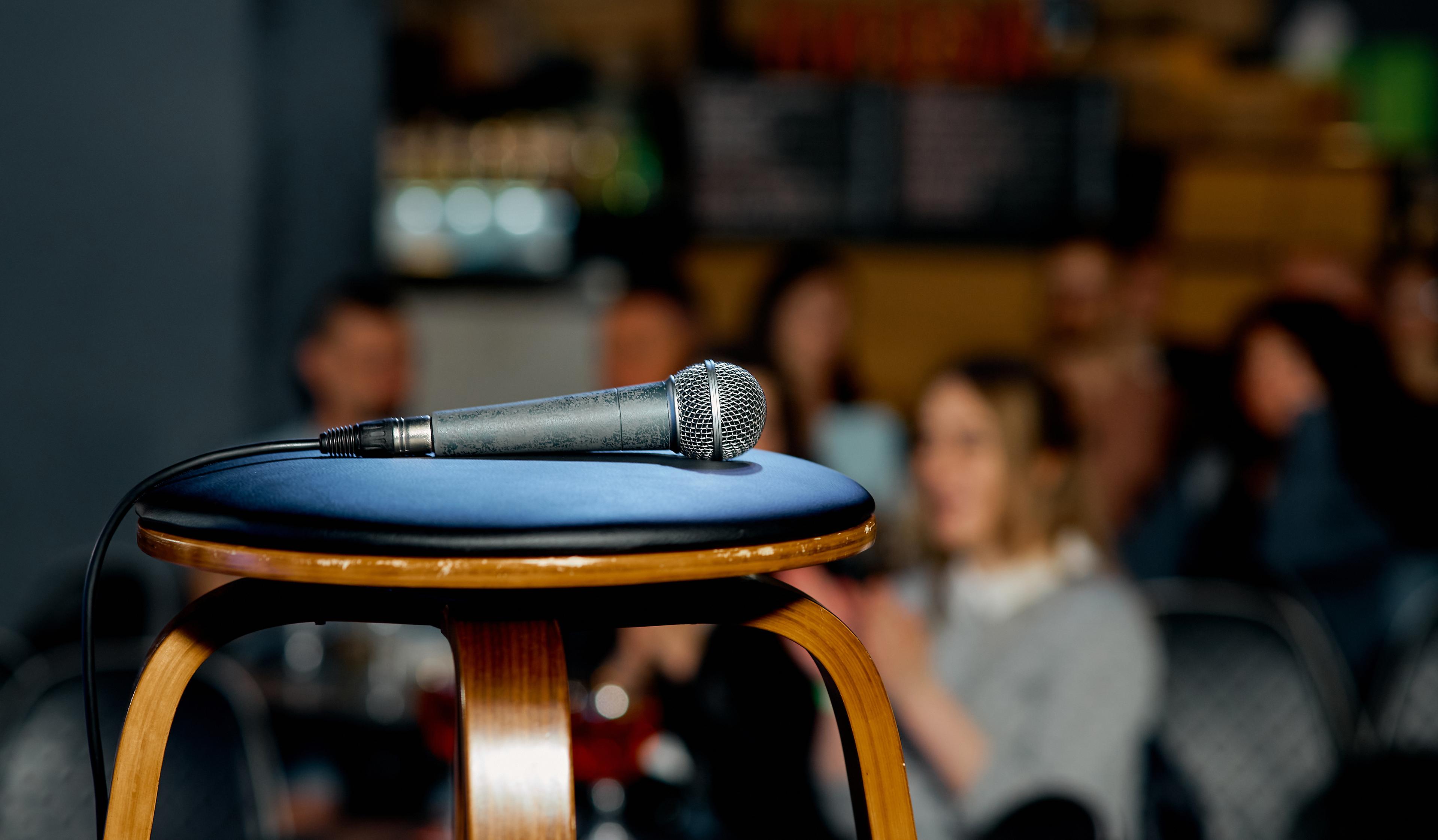 A microphone rests on a stool in a dimly lit venue, with a blurred audience in the background, suggesting an intimate performance setting.