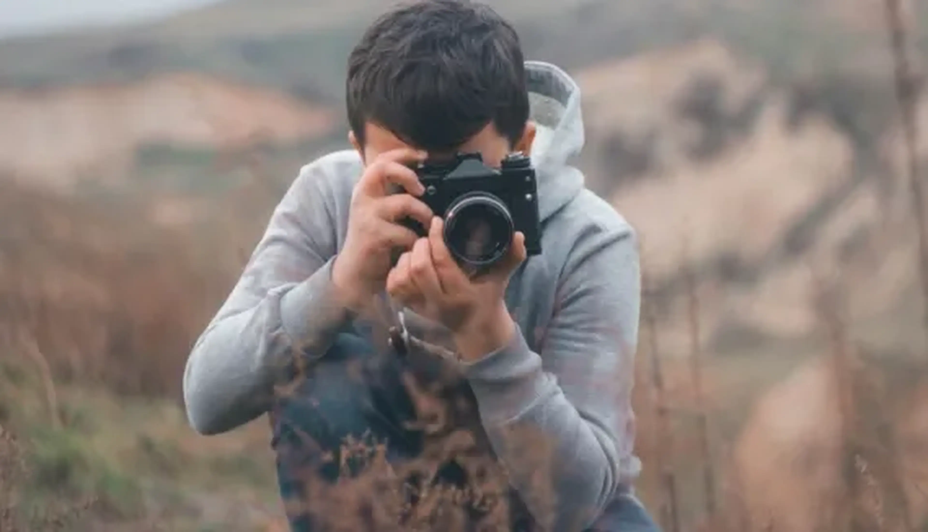Person in a gray hoodie kneeling outdoors, taking a photo with a camera