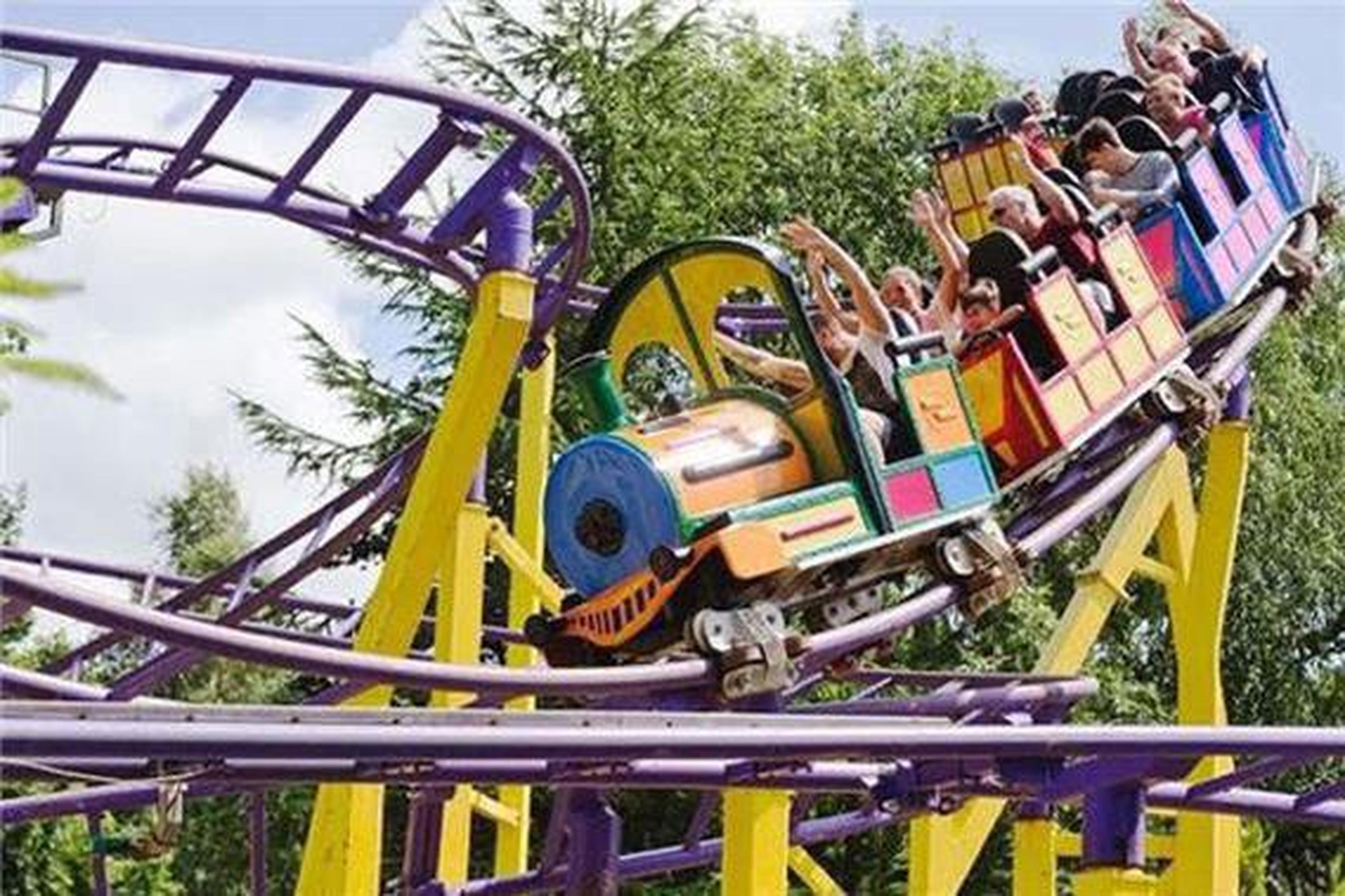 Colorful roller coaster with people raising hands in excitement, surrounded by green trees on a sunny day.
