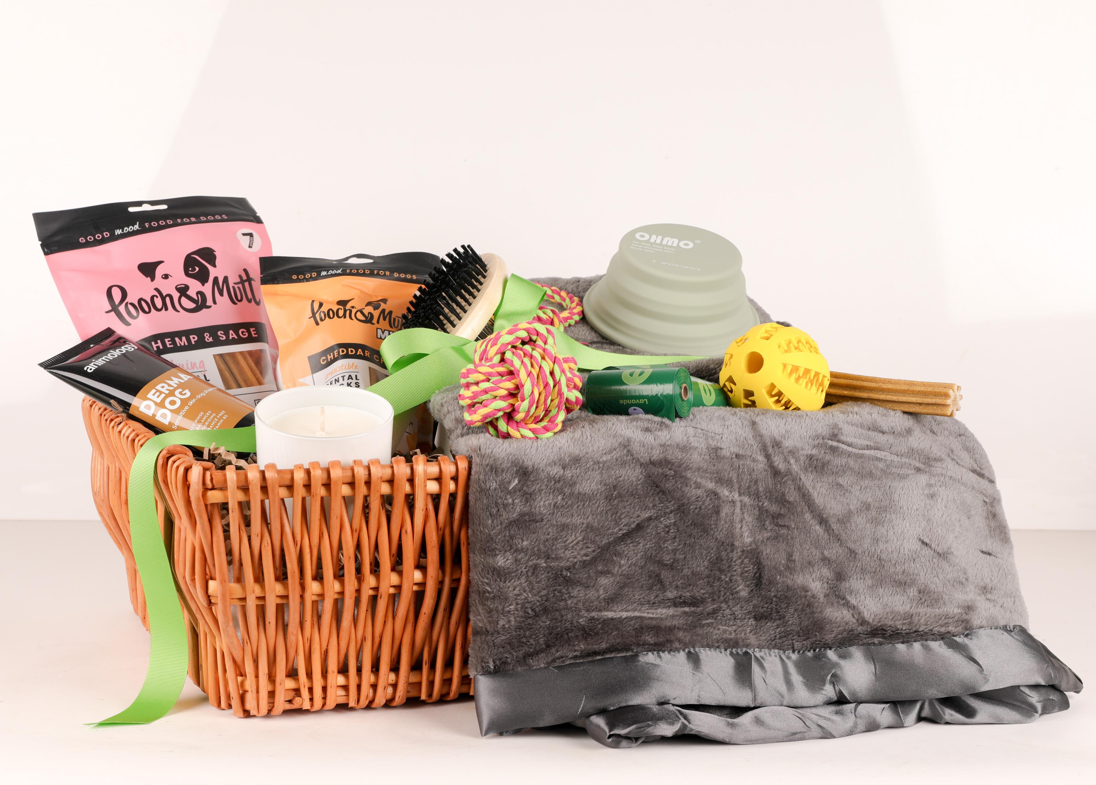 Wicker basket with dog treats, toys, a candle, brush, bowl, blanket, and green ribbon, featuring Pooch & Mutt packaging.