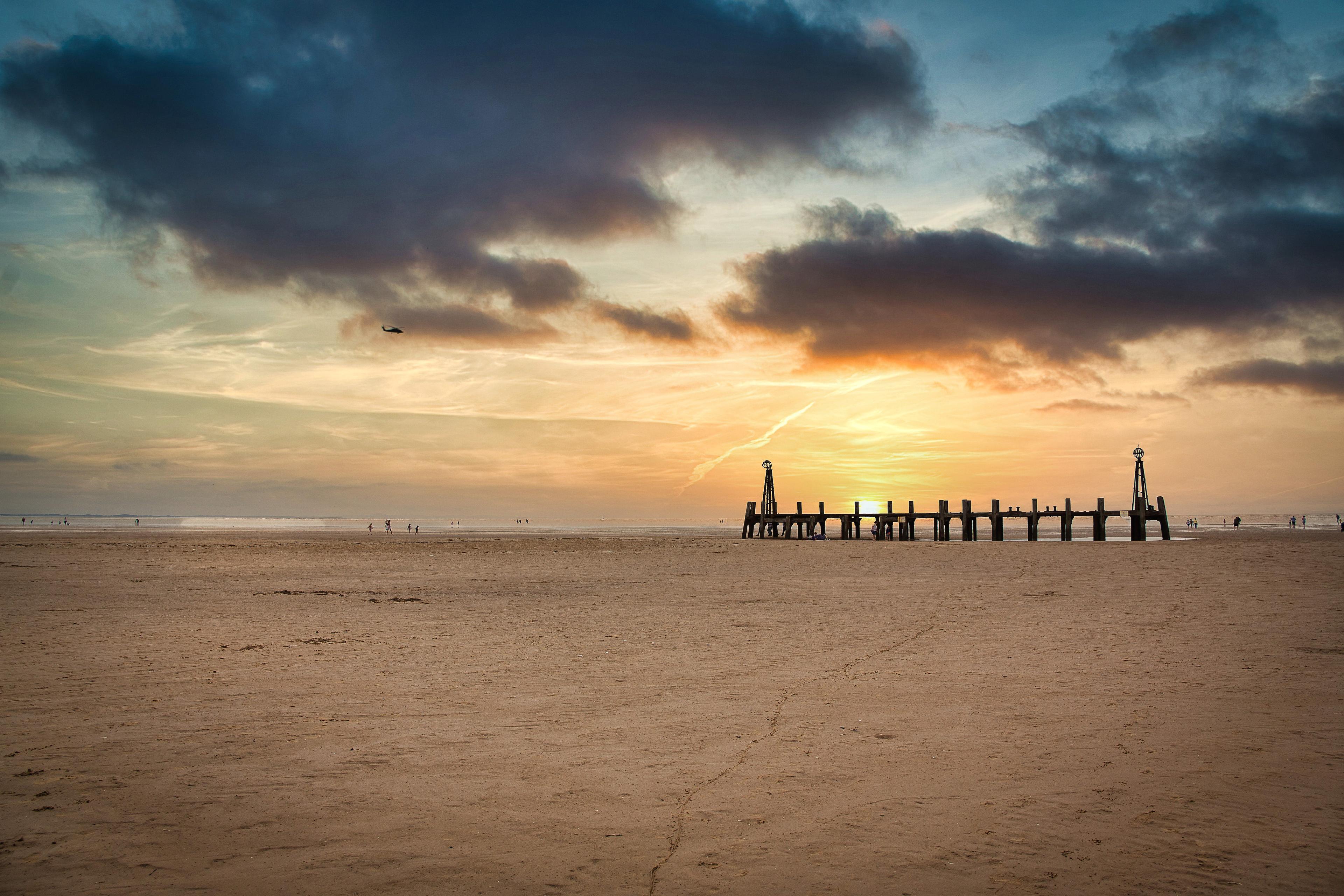 A sandy beach at sunset with scattered people, a wooden pier extending into the sea, and dramatic clouds in the sky.