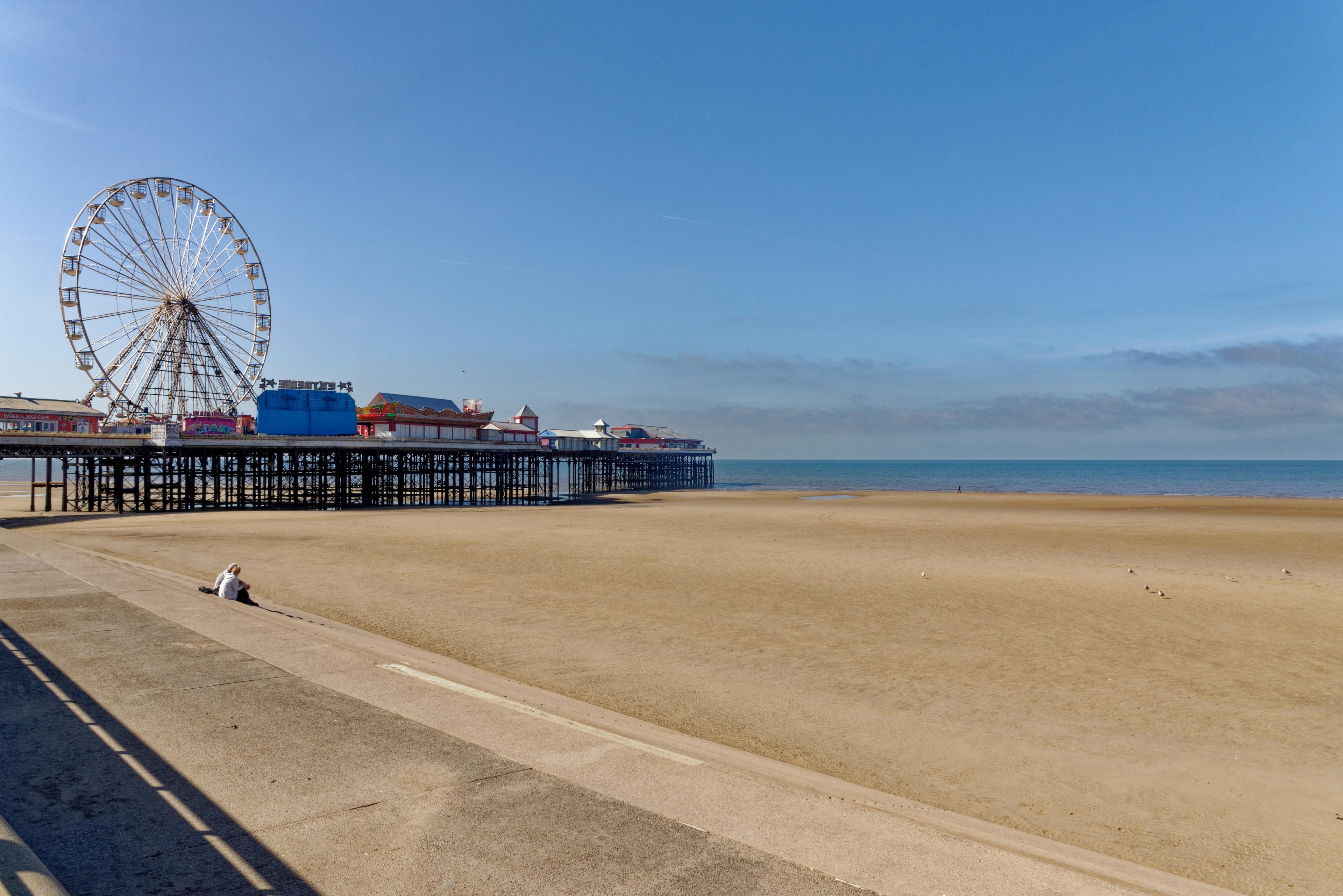 Empty sandy beach with a large Ferris wheel and pier in the background, under a clear blue sky. A person sits on the promenade.